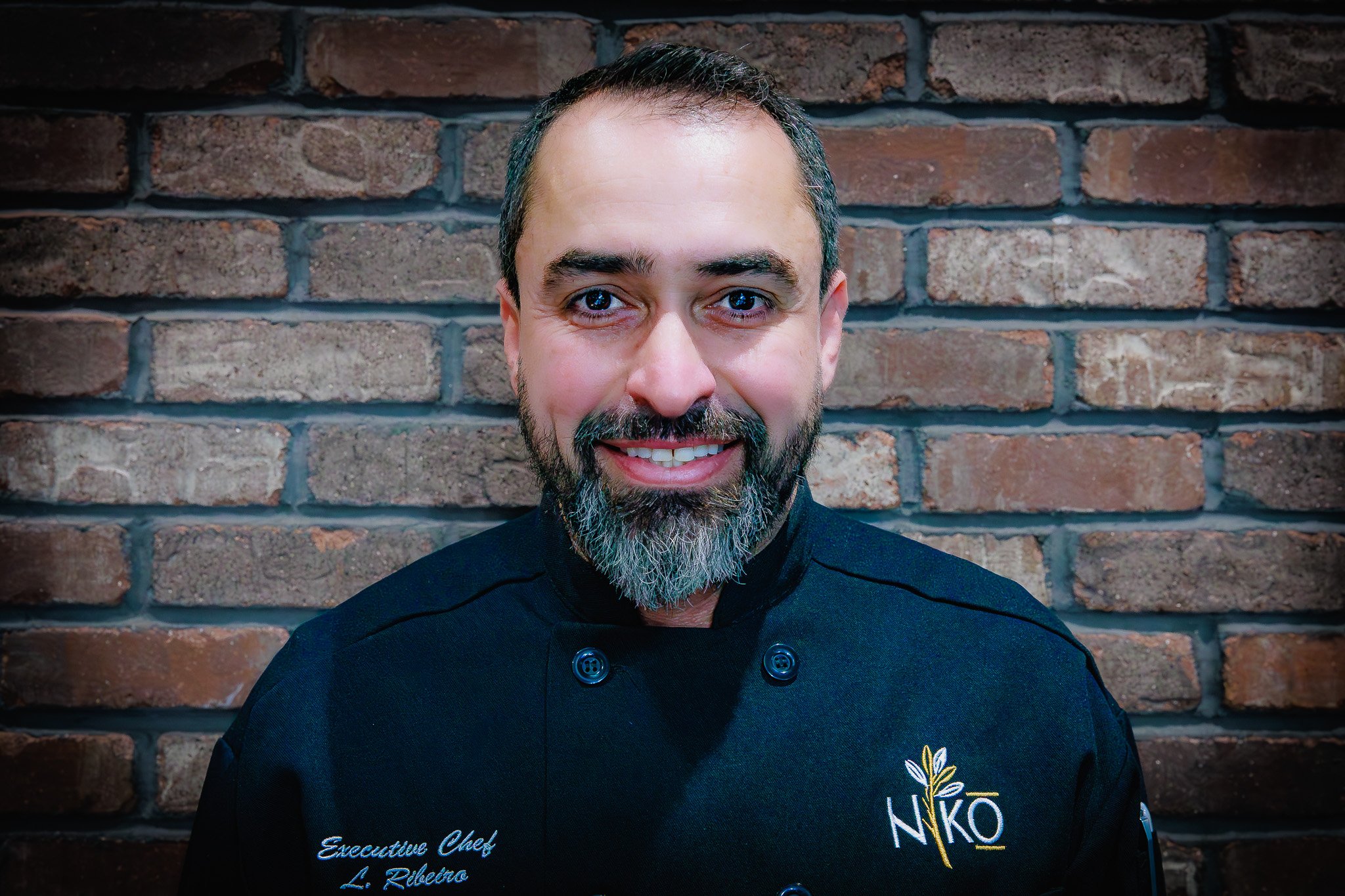 Portrait of a smiling man with a beard, wearing a black chef's jacket, standing against a brick wall.