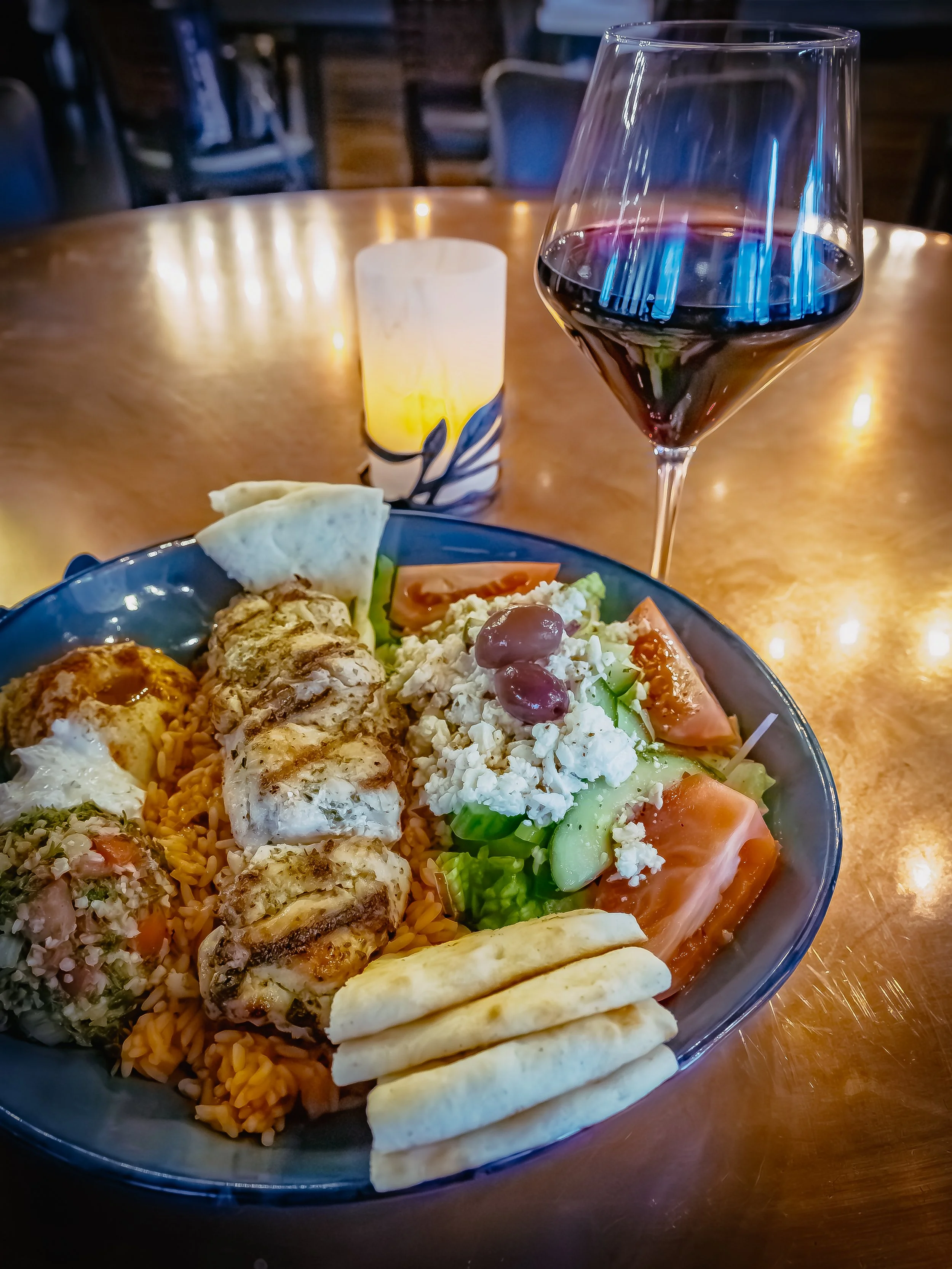 A bowl of Greek salad with tomatoes, cucumbers, feta cheese, and olives, alongside grilled chicken served with rice, and bread slices, with a glass of red wine in the background, lit by a candle on a wooden table.