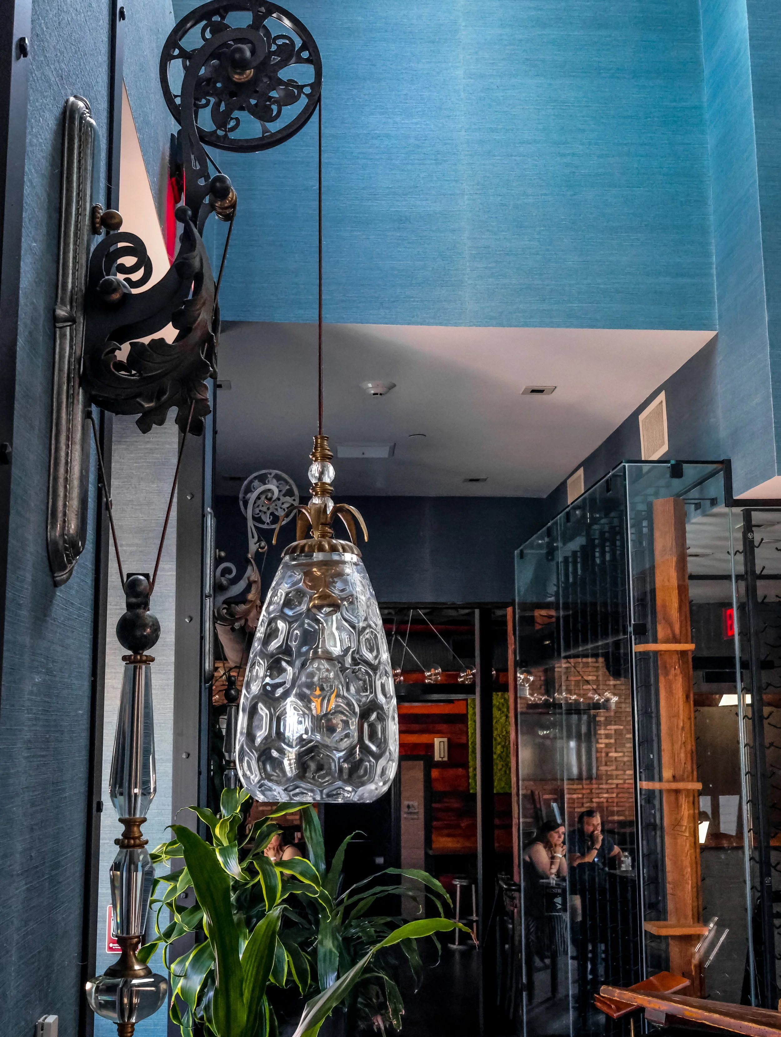 Interior of a restaurant or cafe with blue walls, hanging glass pendant light, decorative wall art, plants, and two women seated at a table in the background.
