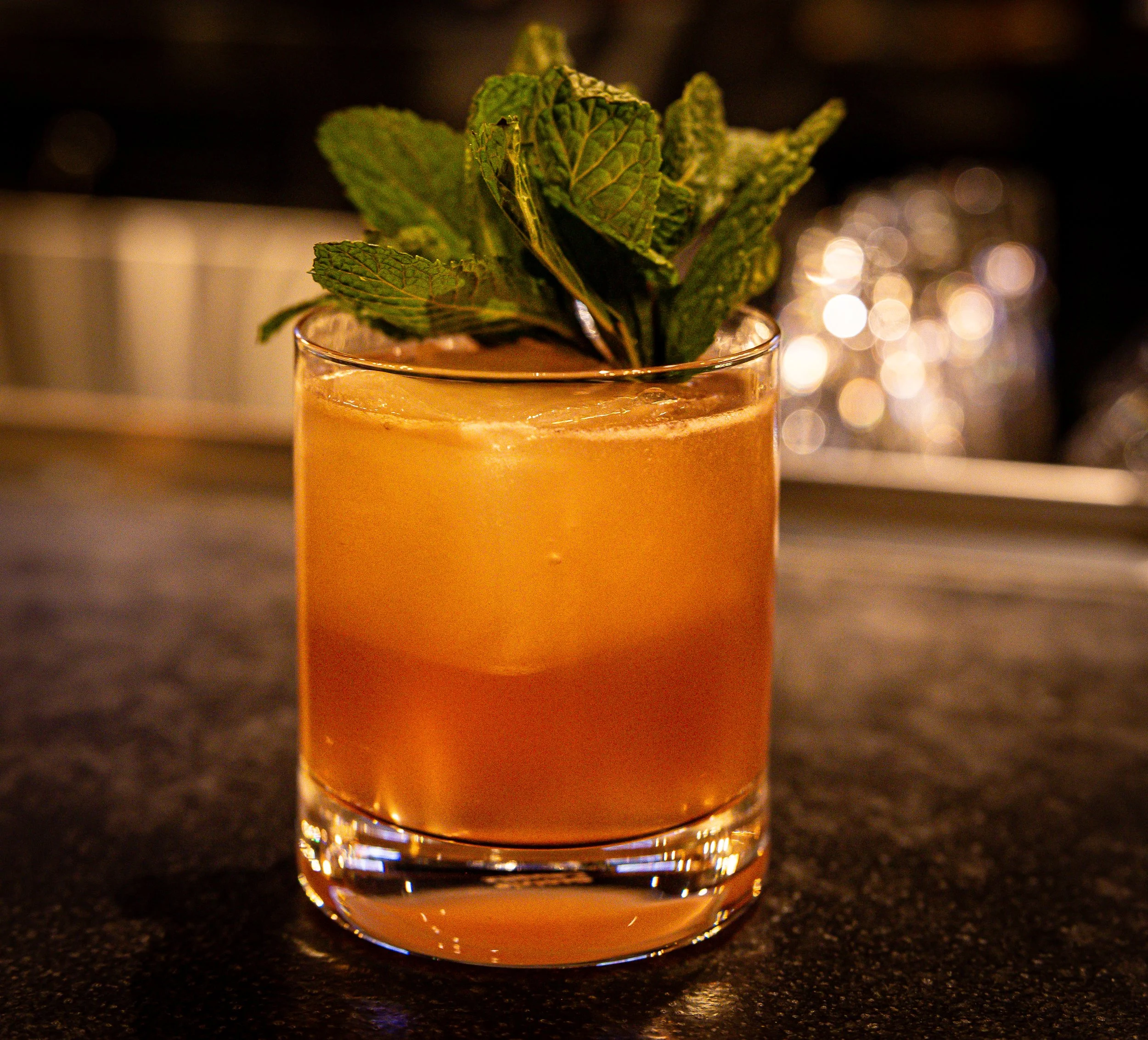 A cocktail with ice and mint leaves in a short glass, on a dark surface with a blurred background.