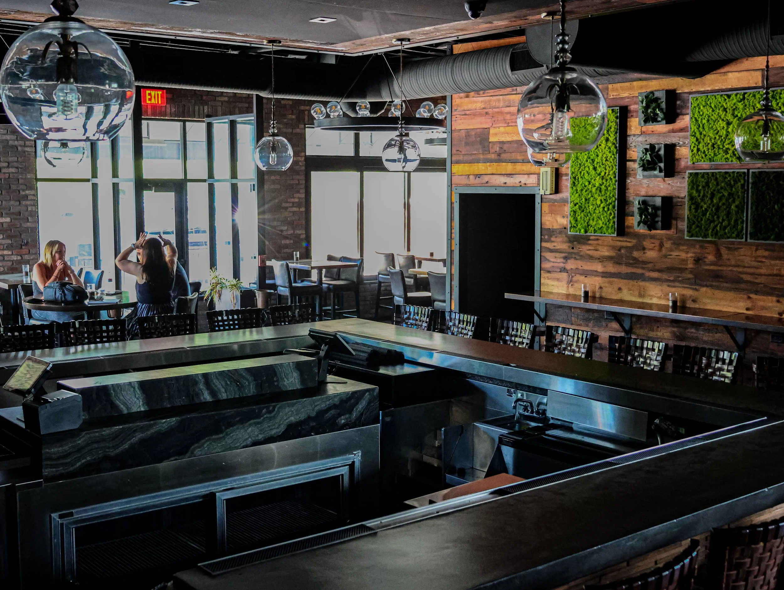 Interior of a modern restaurant with a bar counter, pendant lights, and a wall with green plant decor. Two women are sitting at a table near large windows with sunlight streaming in.