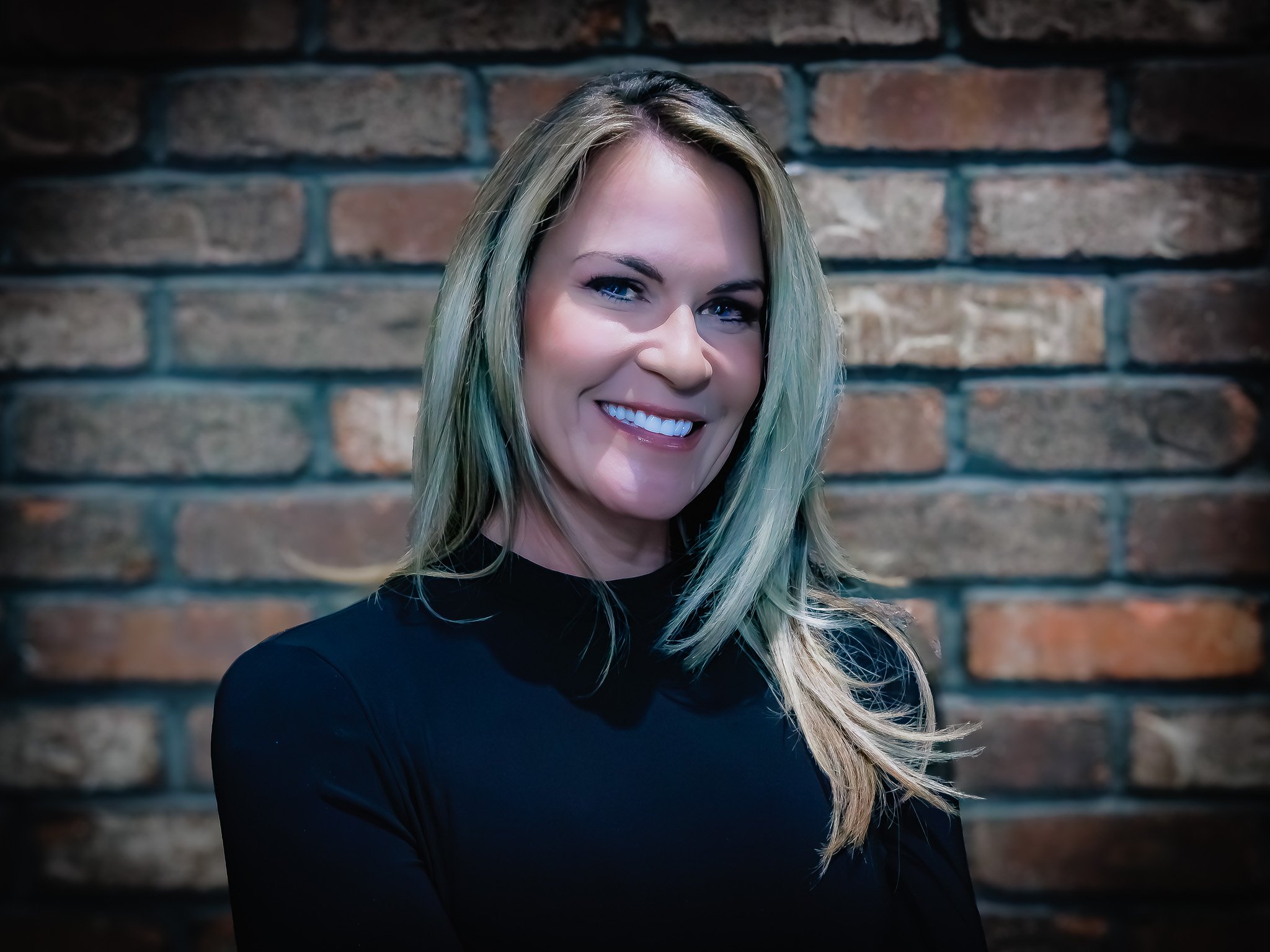 A smiling woman with blonde hair wearing a black top standing in front of a brick wall.