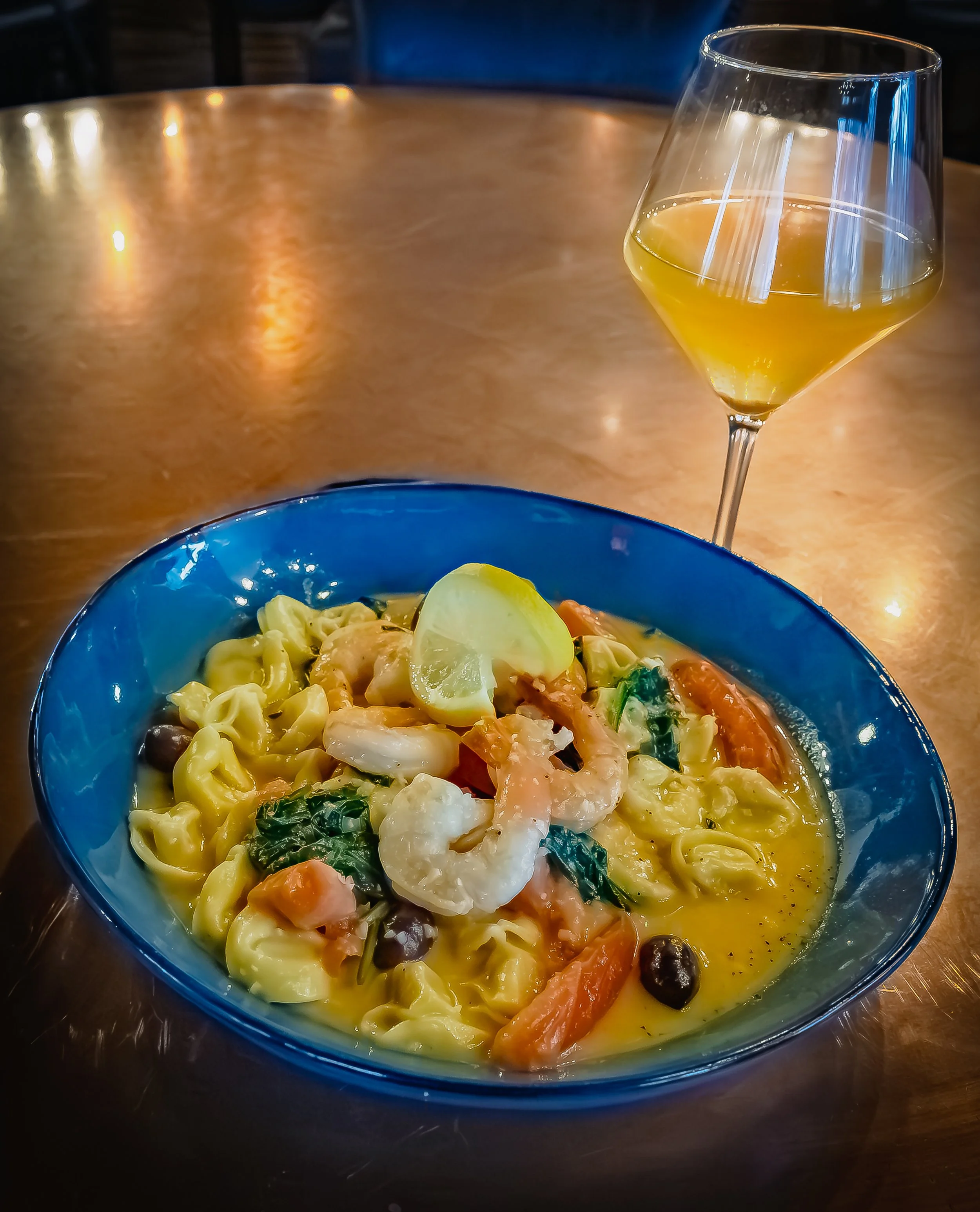 A bowl of seafood pasta with shrimp, vegetables, and lemon wedge, served with a glass of white wine on a wooden table.
