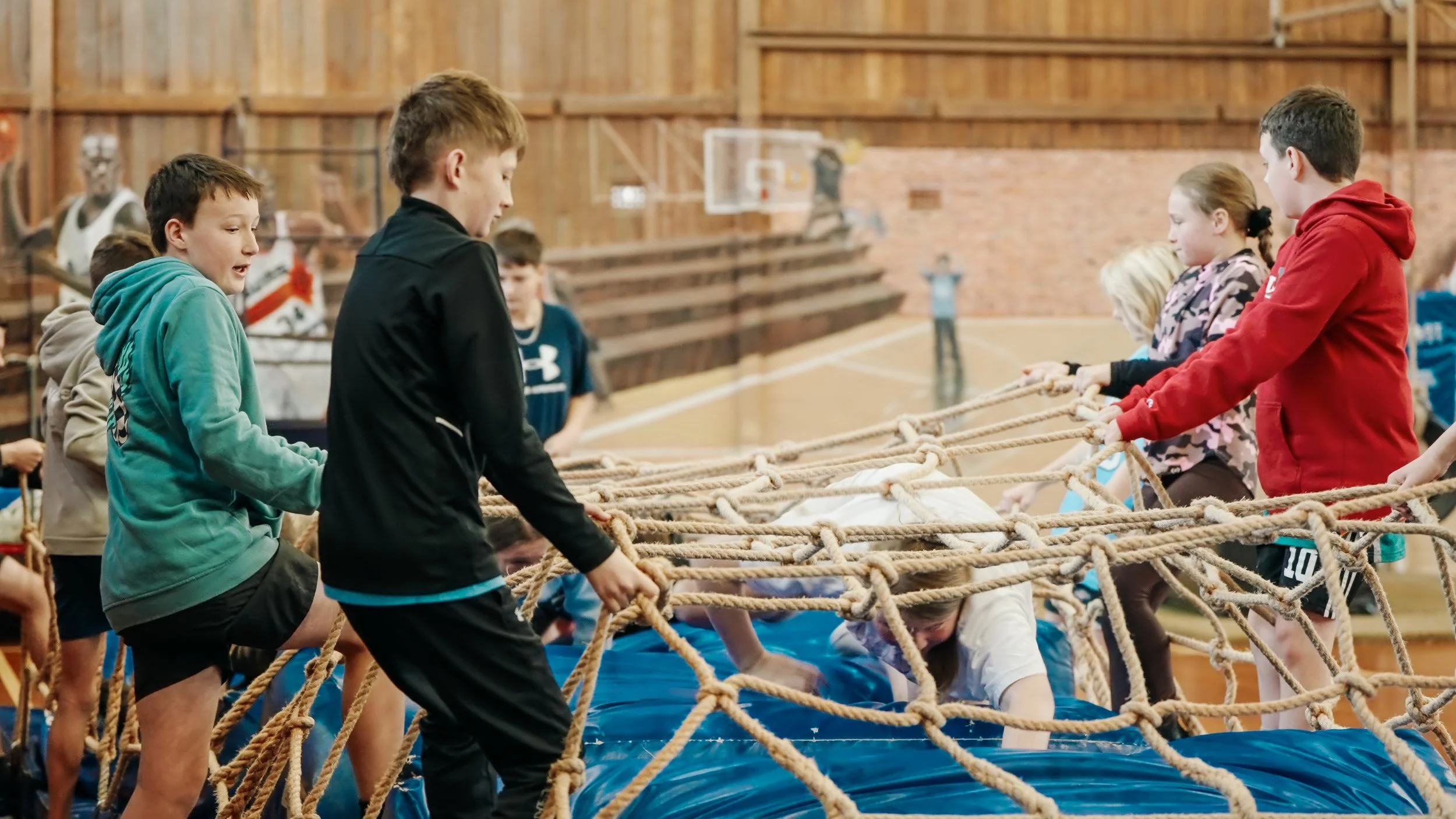 Children participating in an indoor team-building activity involving a large rope obstacle course in a gymnasium.