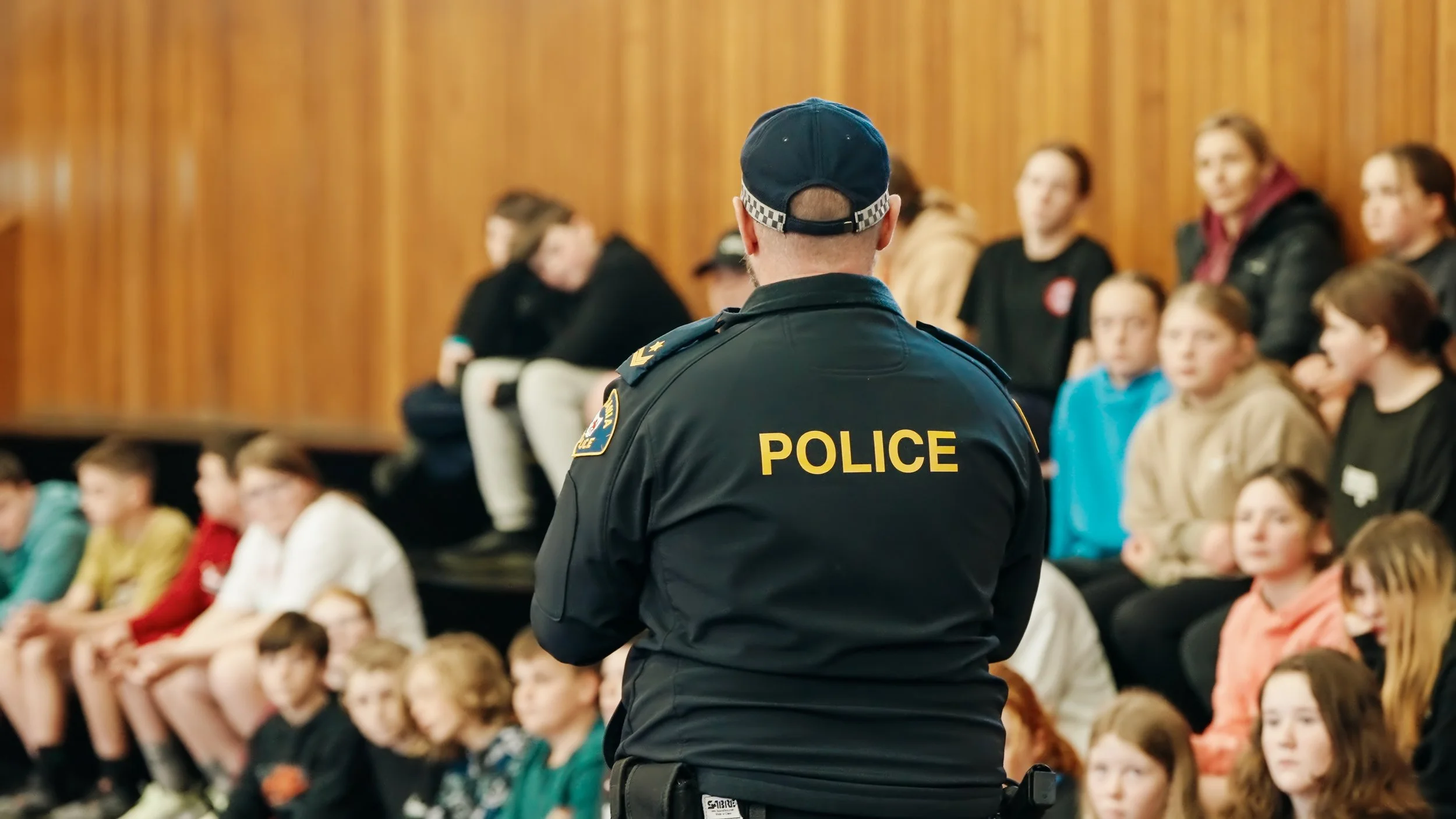 A police officer stands with his back to the camera, facing a group of children and adults seated in a room with wooden walls.