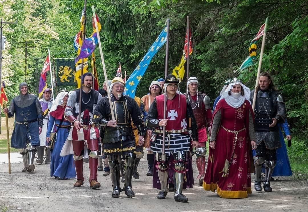 Group of people dressed as medieval knights and nobles, participating in a historical reenactment or parade, walking on a wooded path holding flags and banners.