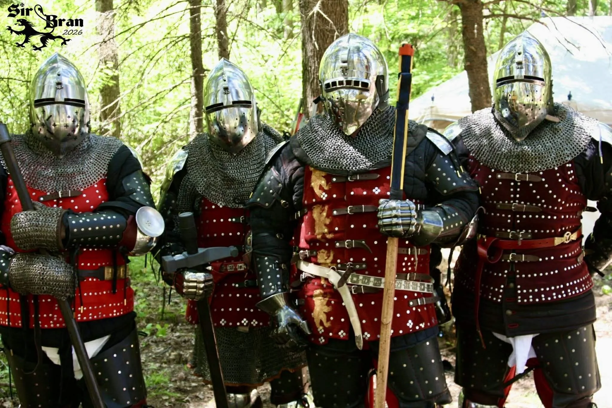 Group of five people dressed as medieval knights in armor and chainmail, standing in a wooded area, with tents in the background.