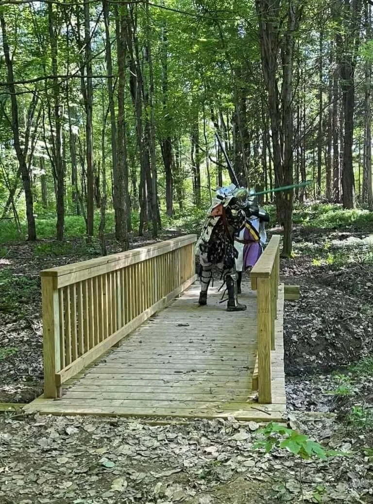 A small wooden bridge in a forest with two people dressed in costumes, one of whom has a sword.