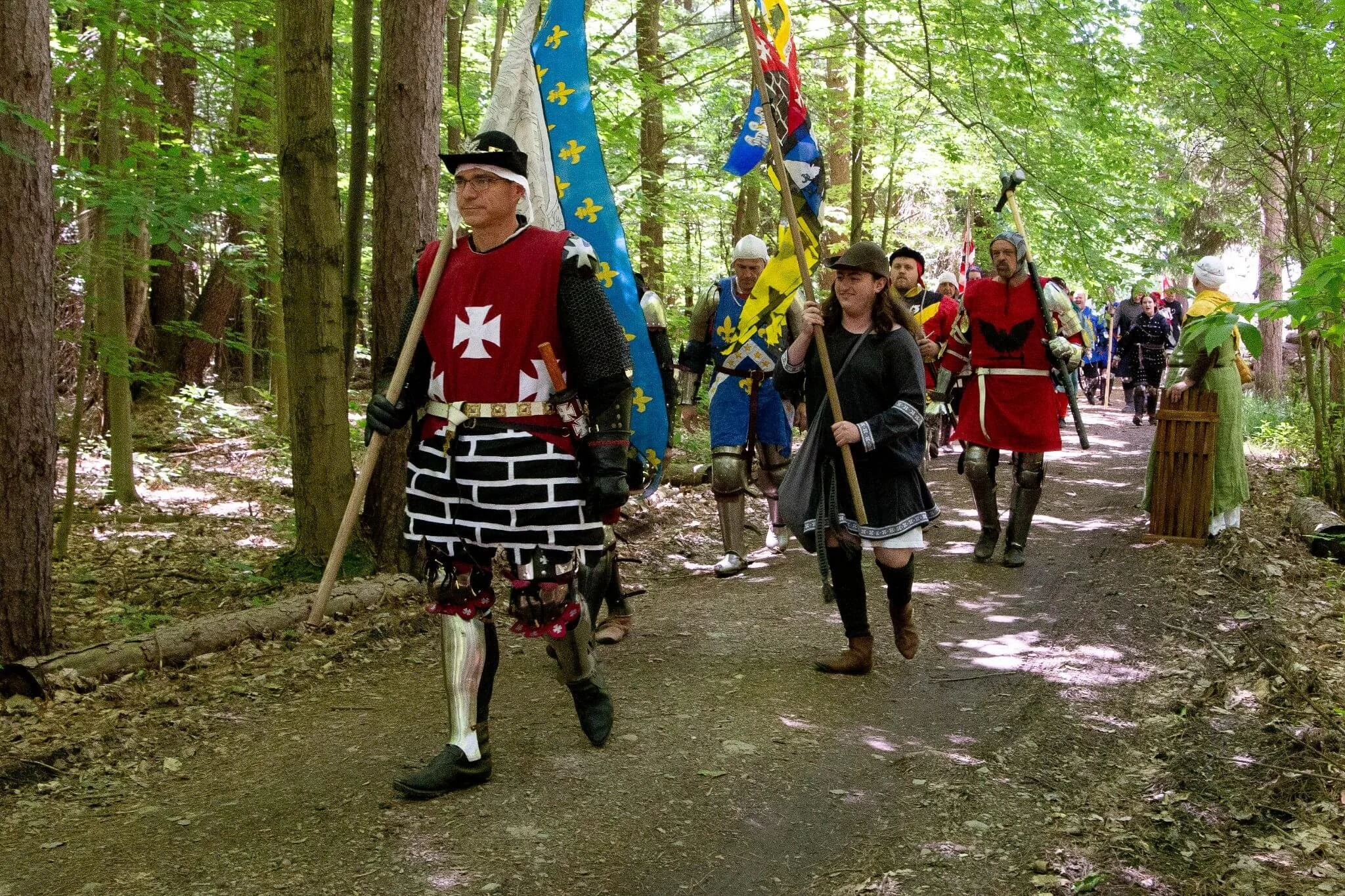 People dressed in medieval costumes and armor, carrying flags, walking through a wooded area during a historical reenactment event.
