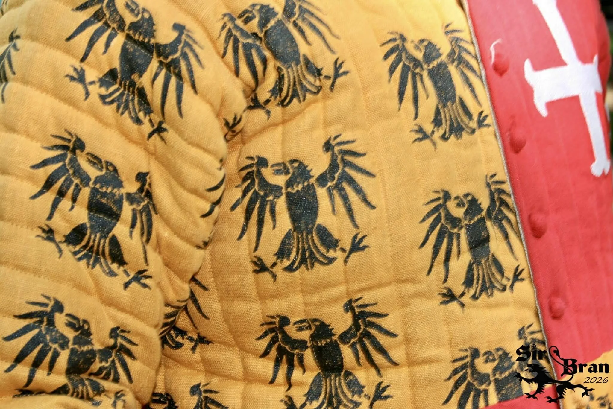 A yellow quilted fabric with black lobster patterns, next to a red fabric with white cross stitching and a logo in the bottom right corner reading 'Sir Bran 2026'.