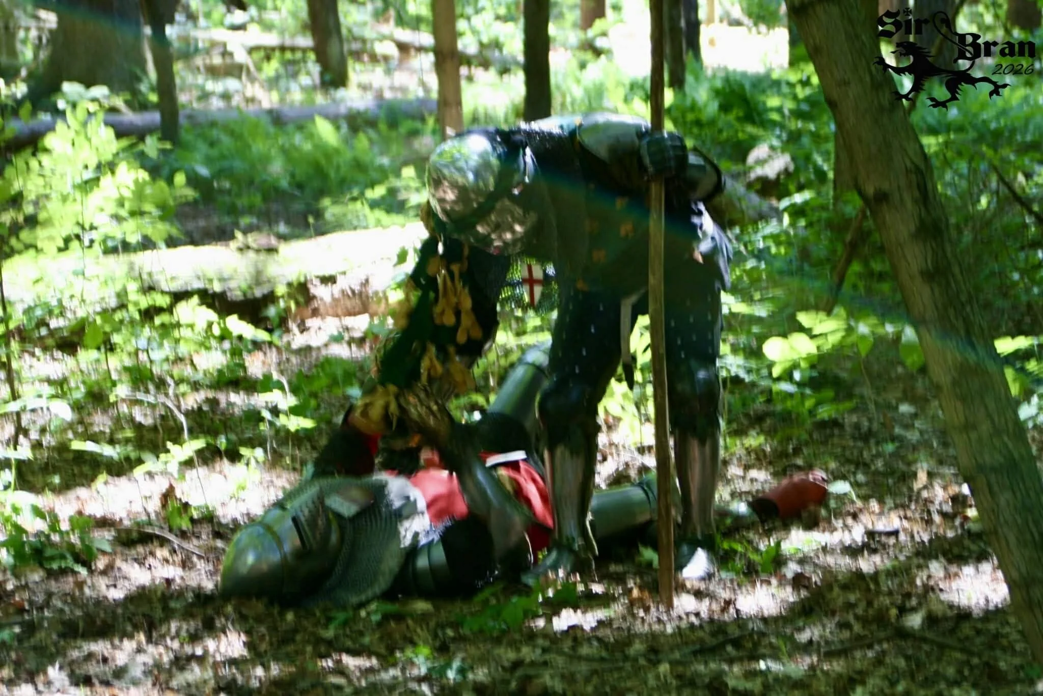 A humanoid robot with a black and metallic body bending over in a forested area, appearing to assist or interact with a person lying on the ground. The robot has a stick in its right hand, and the person on the ground is dressed in casual clothing, w
