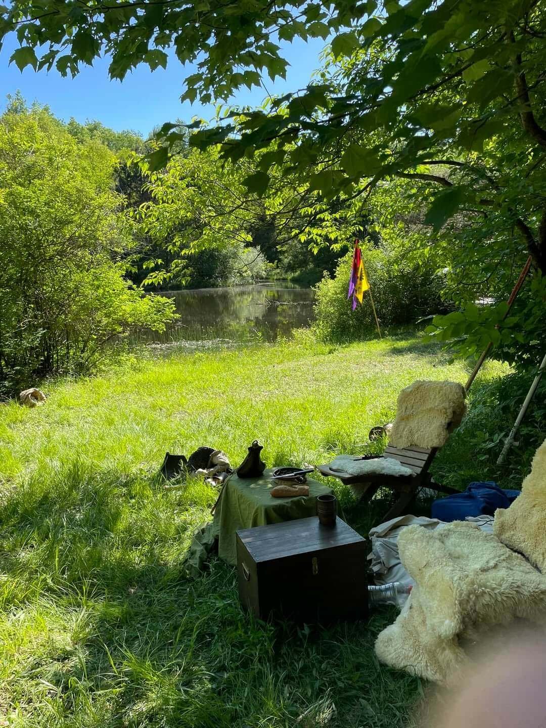 A peaceful riverside scene with green trees, grass, and a calm river. There is a small makeshift outdoor seating area with a lounge chair covered in a fluffy white throw, a small table with various items, and colorful flags. The scene is bright and sunny.