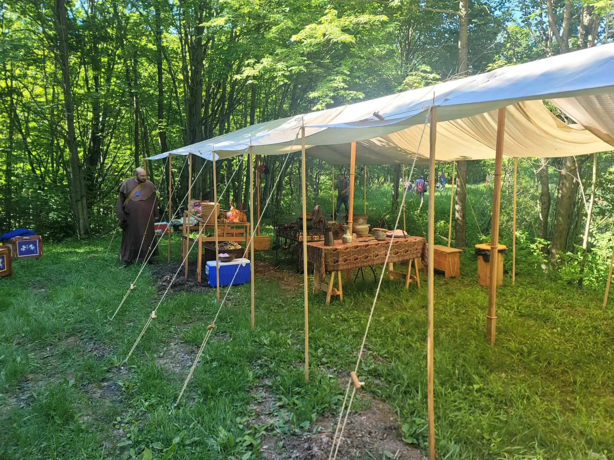 Outdoor market stall set up under a canopy in a wooded area, with tables displaying various items, including pottery and artwork, and a monk or man in robes nearby.