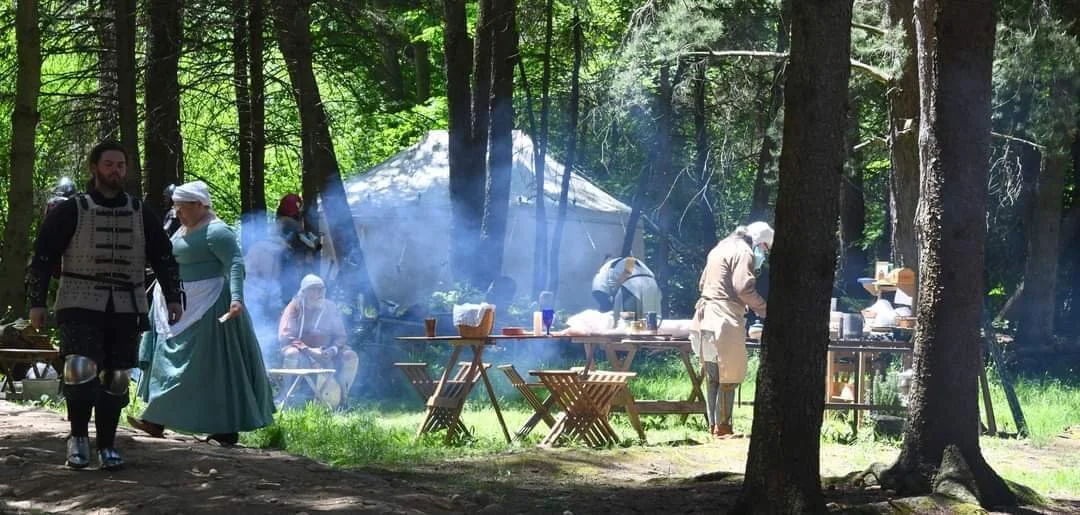 Historical reenactment in a forest with people dressed in medieval clothing, cooking around a campfire, with tents and wooden tables.