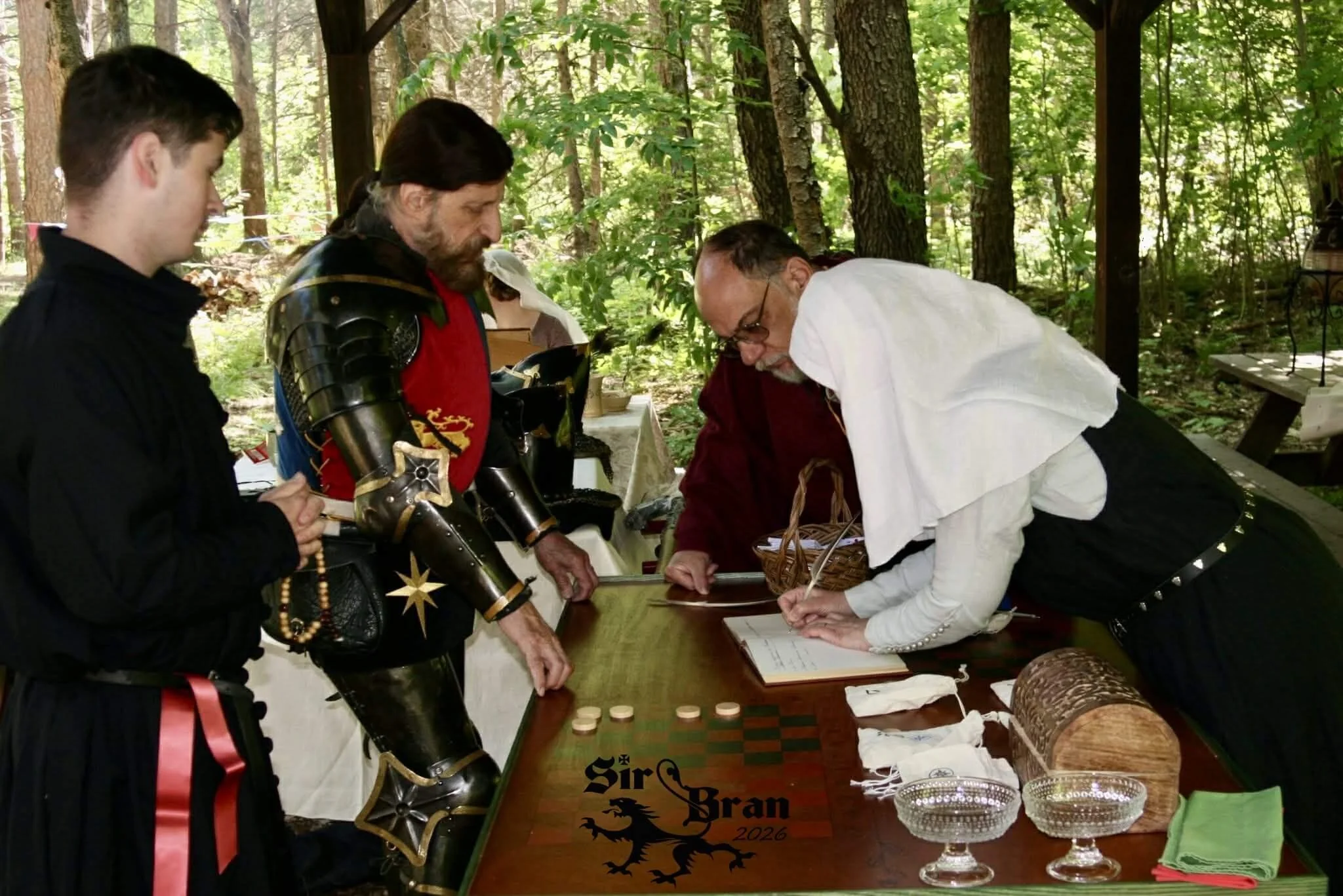People in medieval and renaissance costumes gather around a table outdoors in a wooded area, engaging in a reenactment or event. The table has various items, including a chessboard, a chest, glass bowls, and cloth. One man dressed as a knight is hold