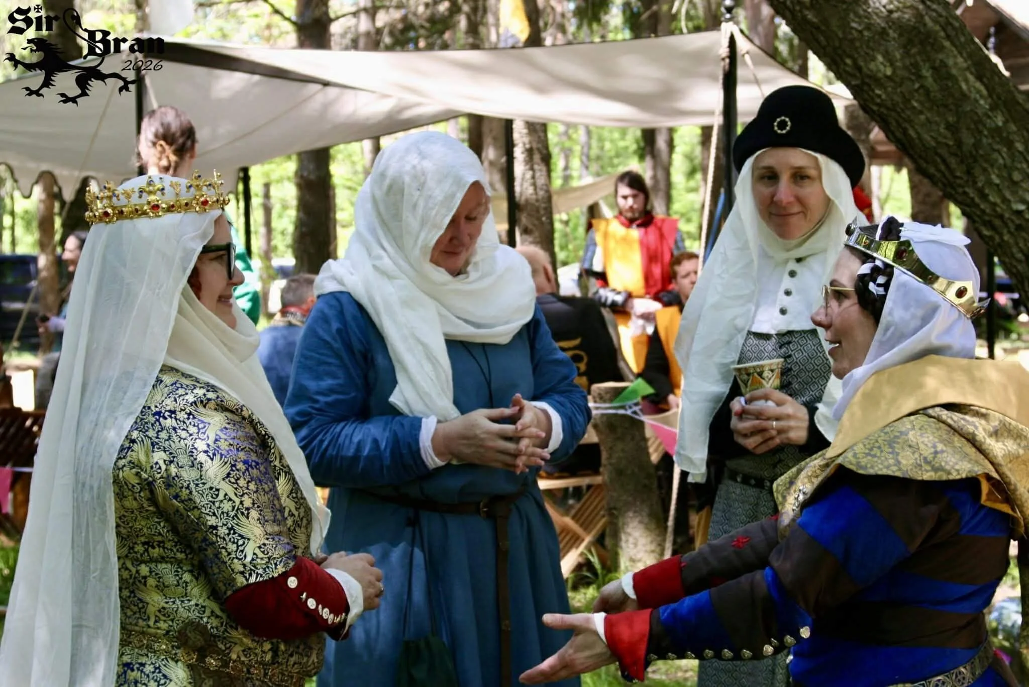 Four women in medieval costumes with crowns and head coverings, engaged in conversation at an outdoor event in a wooded area, with other people and tents in the background.