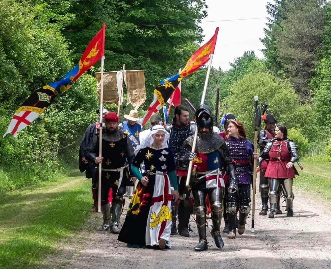 Group of people dressed in medieval armor and costumes, carrying flags and walking down a wooded path.