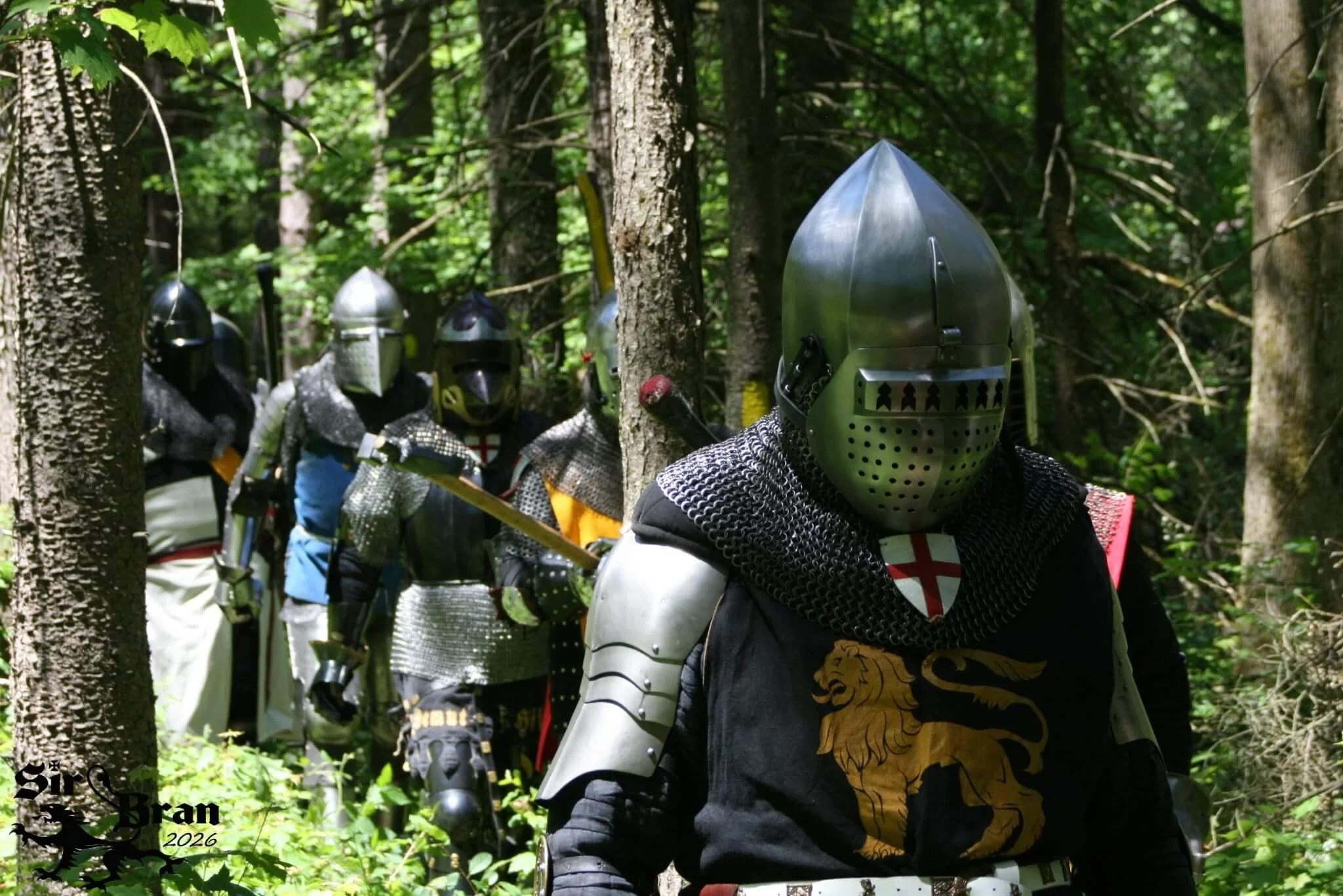 A group of people dressed as medieval knights wearing helmets and armor, walking in a dense forest.