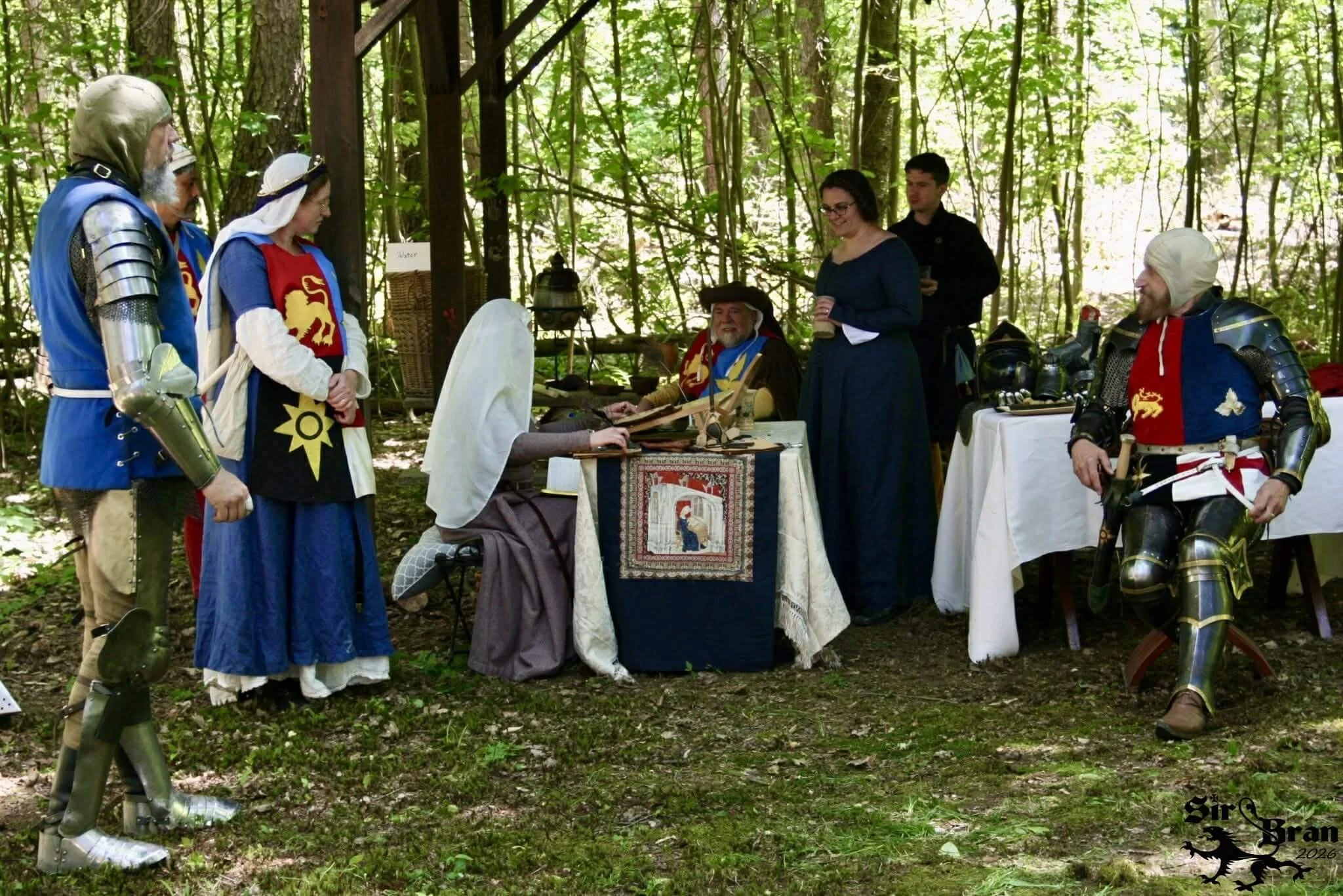 Reenactment of medieval knights and characters in costume, gathered outdoors in a wooded area, with a table displaying medieval artifacts.