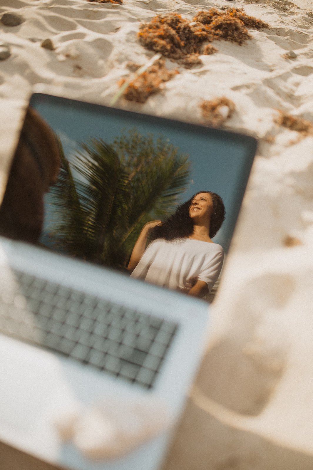 Laptop en la arena con reflejo de mujer y palmera en fondo tropical.
