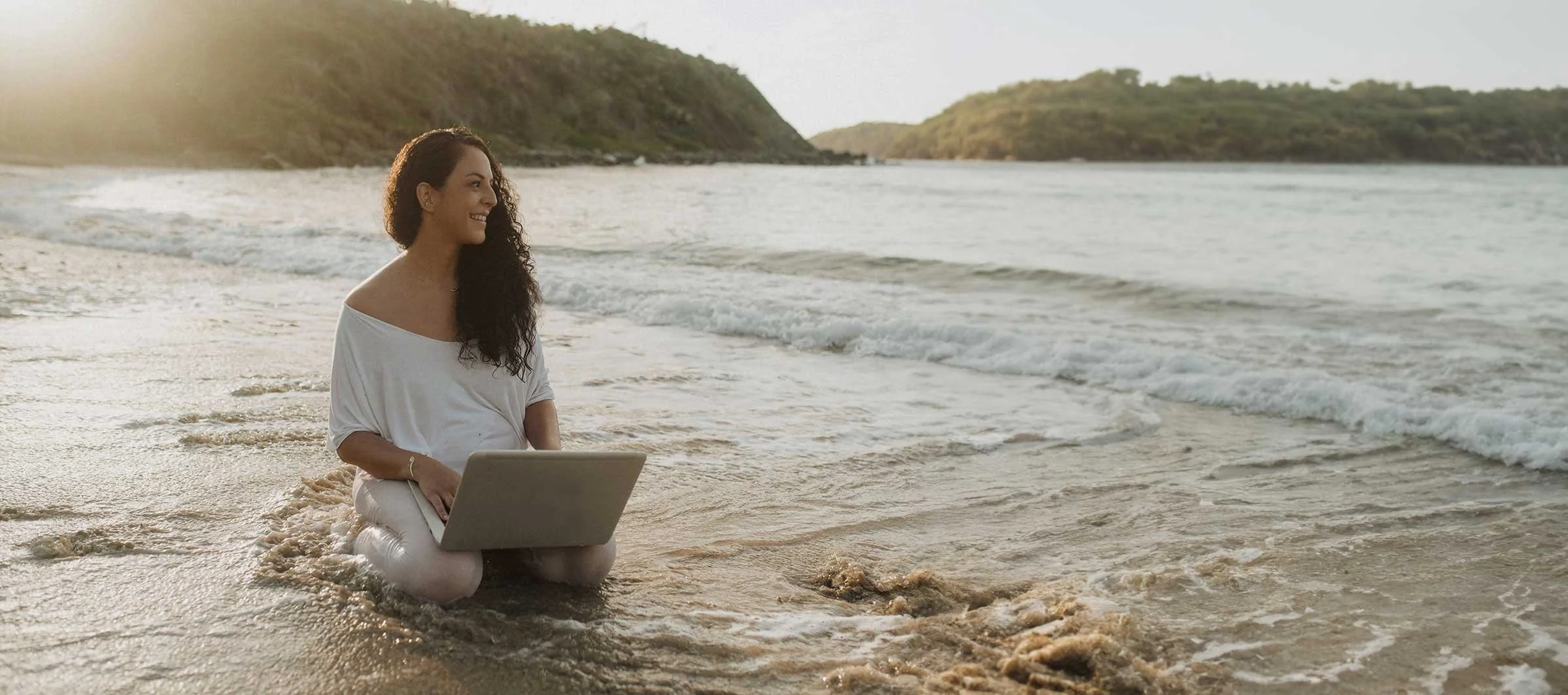 Mujer sonriente con laptop en la orilla del mar, luz cálida y olas suaves.