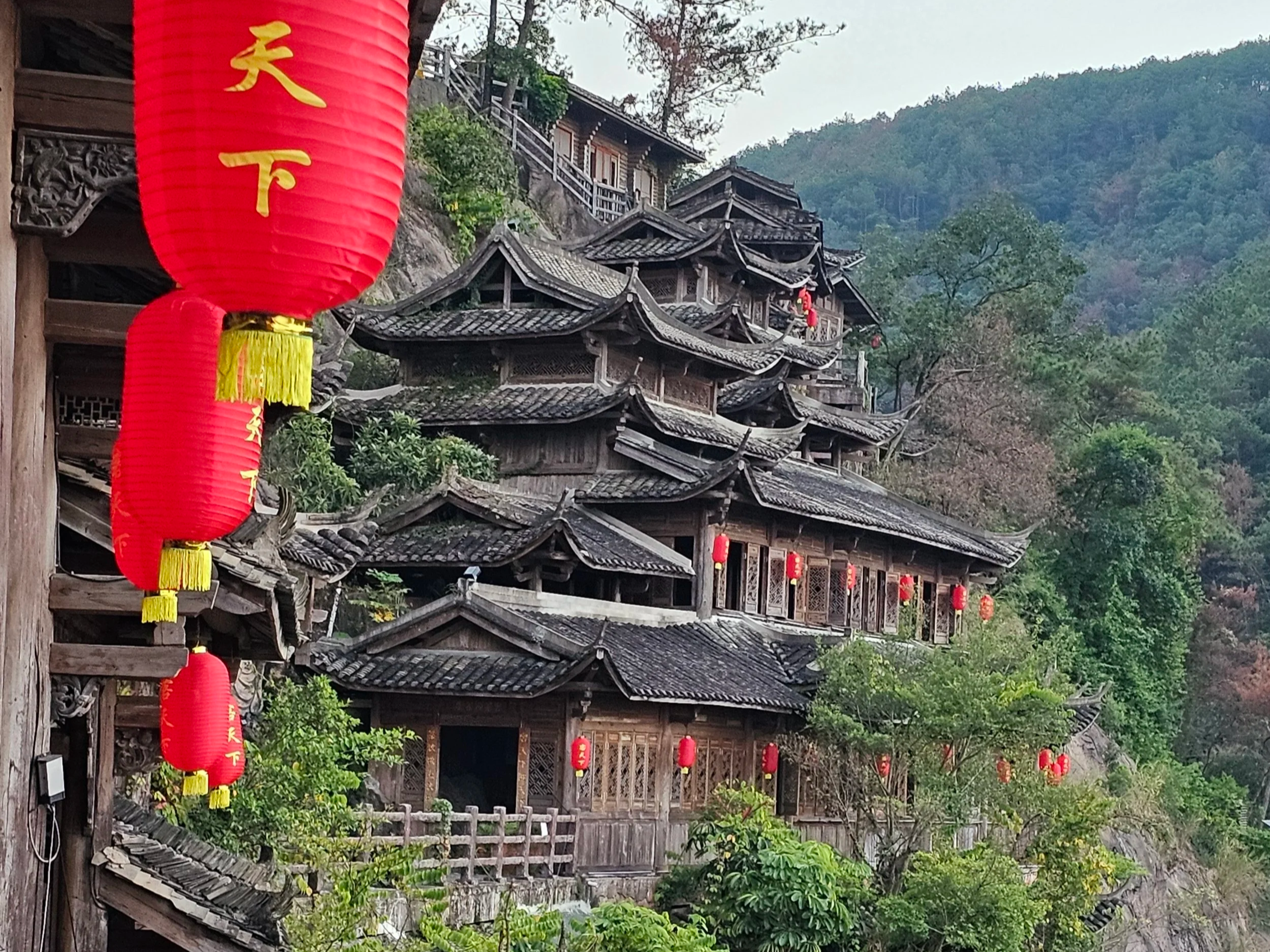 Traditional multi-tiered wooden pagoda with curved roofs on a hillside, decorated with red lanterns, surrounded by lush green trees and mountains in the background.