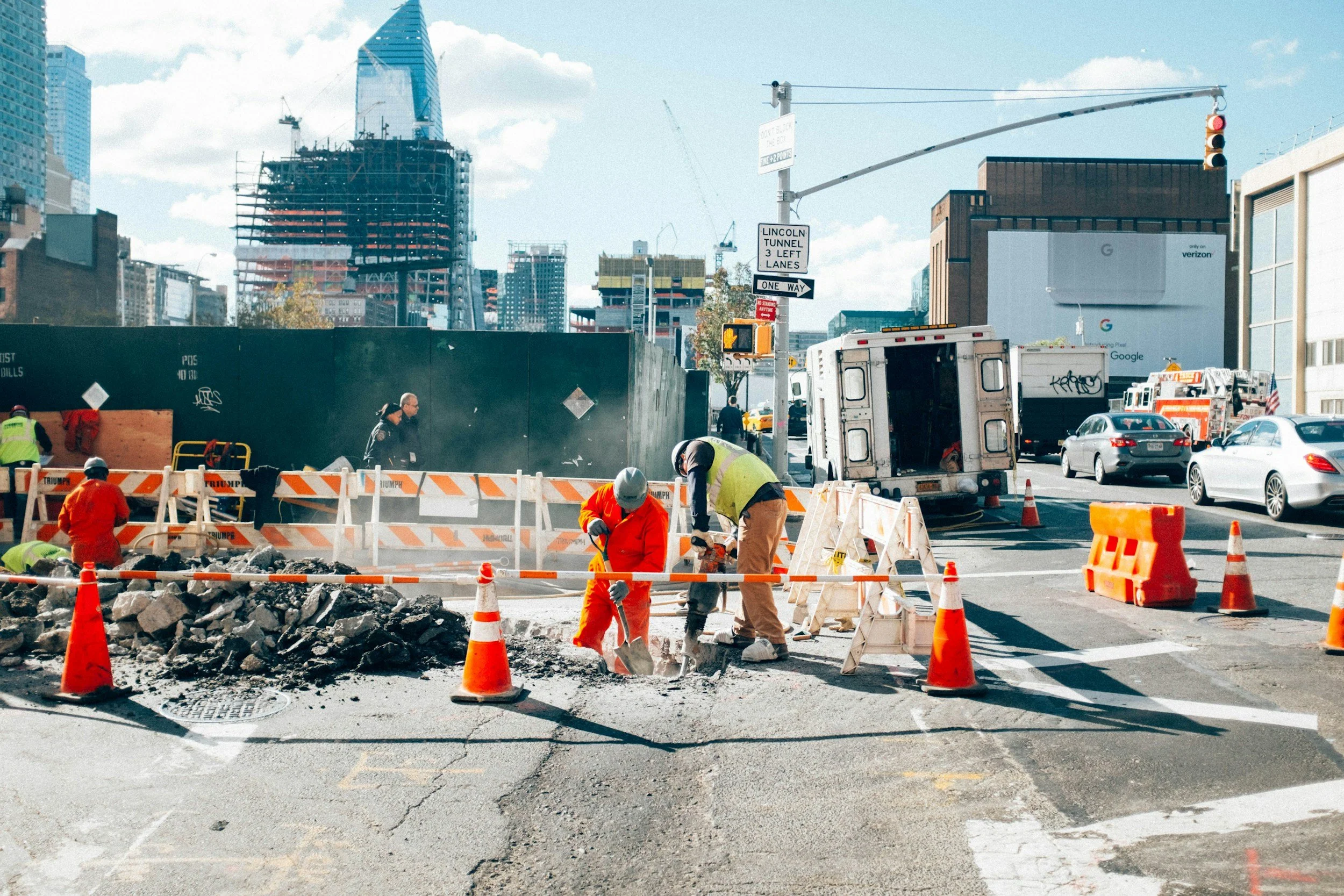 Construction workers in safety gear working on a city street with orange cones and barriers, vehicles, and tall buildings in the background.