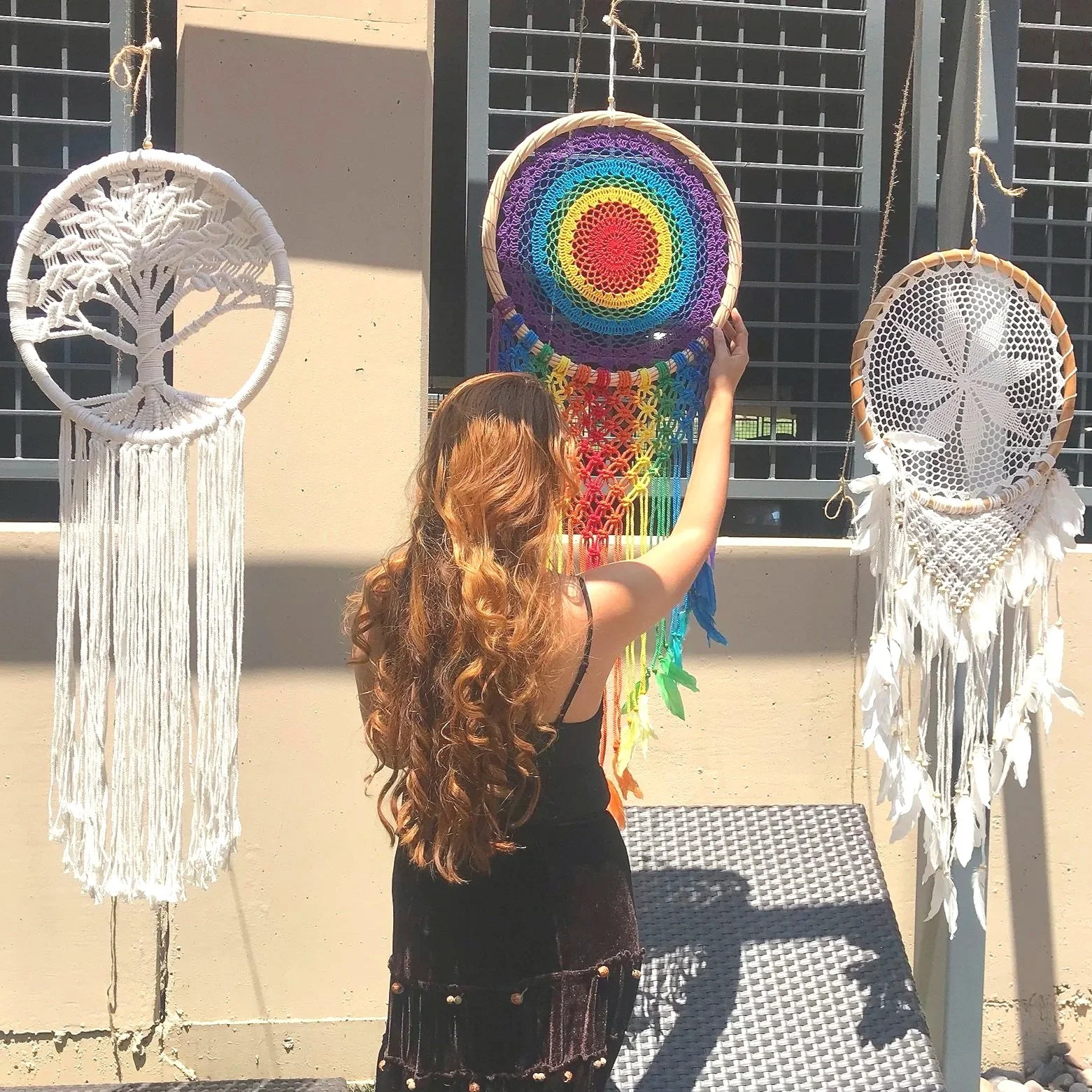 A woman with long, wavy, light brown hair adjusts a colorful rainbow dreamcatcher display. Two other white dreamcatchers are hanging on either side. The scene is outdoors with a beige wall and metal window grilles in the background.