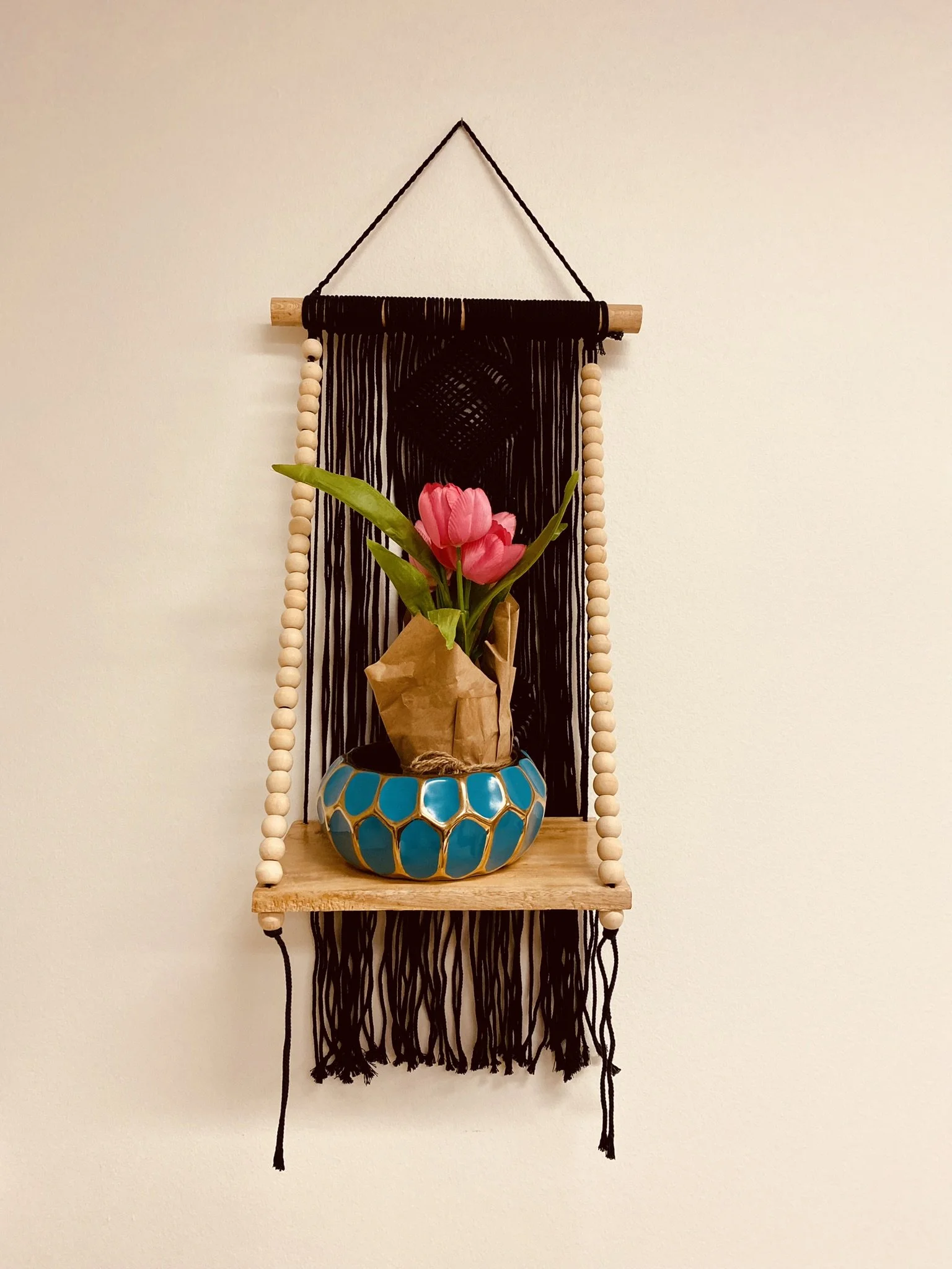 Decorative hanging wall piece with black string, a wooden rod, and black tassels, featuring a small wooden shelf holding a turquoise and gold vase with pink tulips wrapped in brown paper.