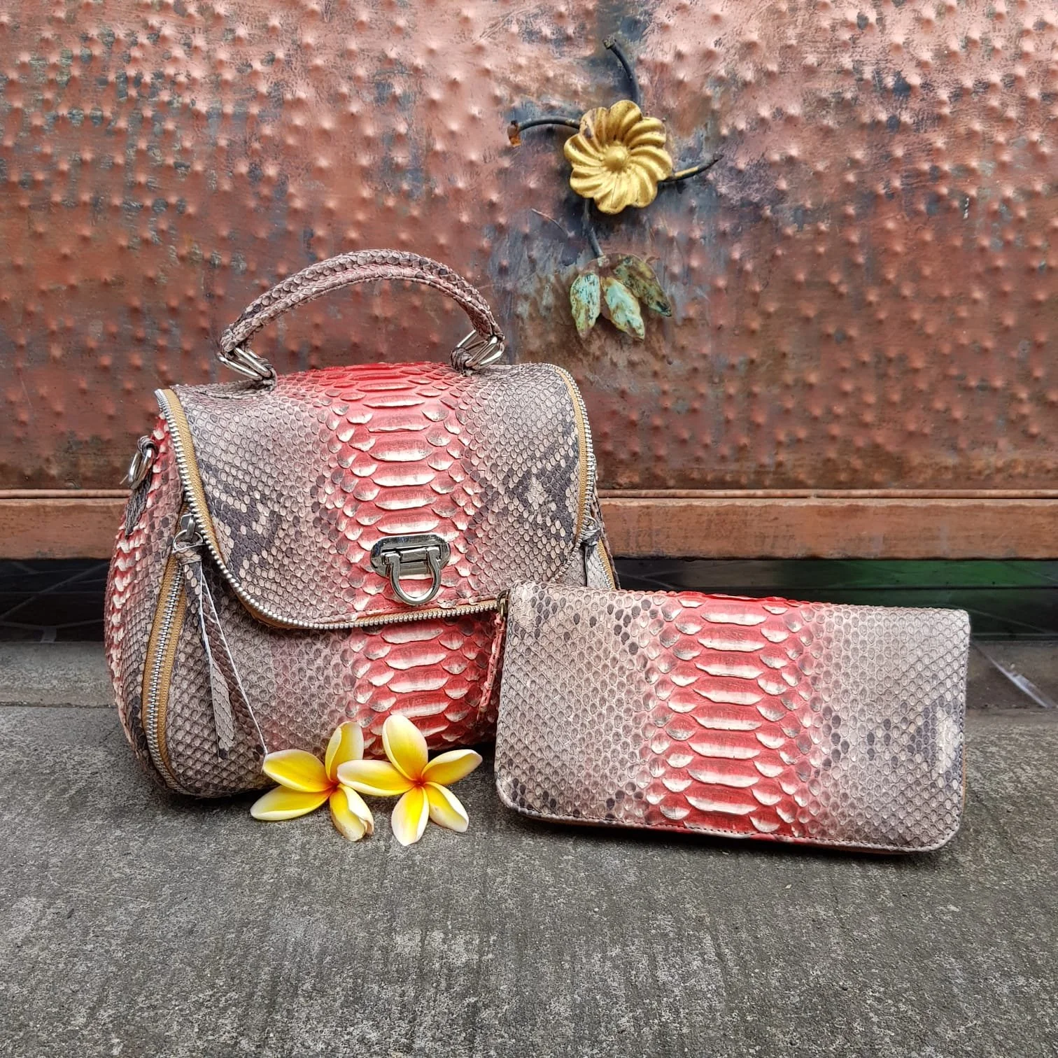 A handbag and wallet with a snakeskin pattern in pink, beige, and black, placed on a concrete surface. There are yellow and white flowers and a yellow flower-shaped wall decoration in the background.