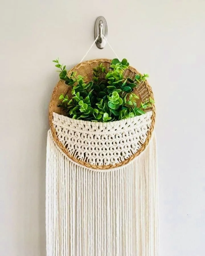 Wicker wall hanging with green plants, white crochet, and long white yarn on a white wall.