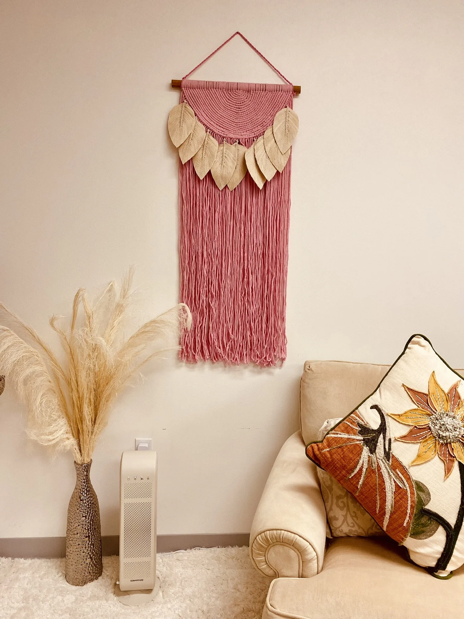 Decorative wall hanging with pink string, beige leaves, and a wooden dowel, hanging above a beige sofa with a floral pillow, next to a vase with pampas grass and a white air purifier.