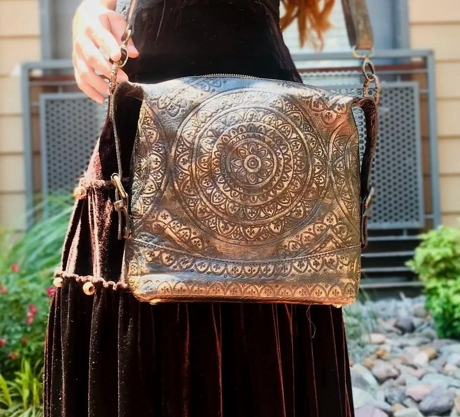 Person holding a handbag with intricate circular patterns, standing outdoors near a building with a wooden exterior and rocks in the garden.