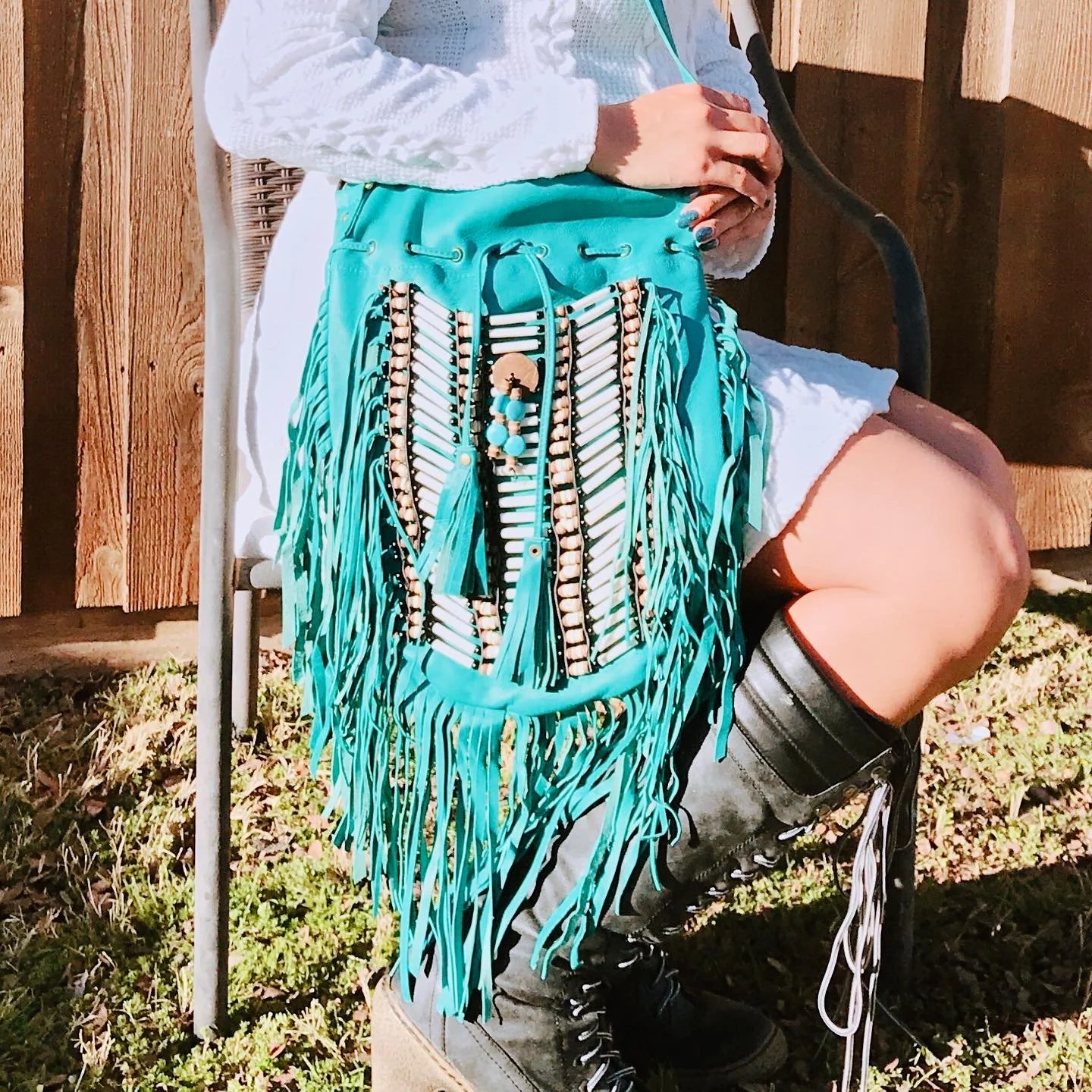 Person sitting outdoors on a chair, wearing black boots, shorts, and a white top, holding a large turquoise bag with fringes and beads.