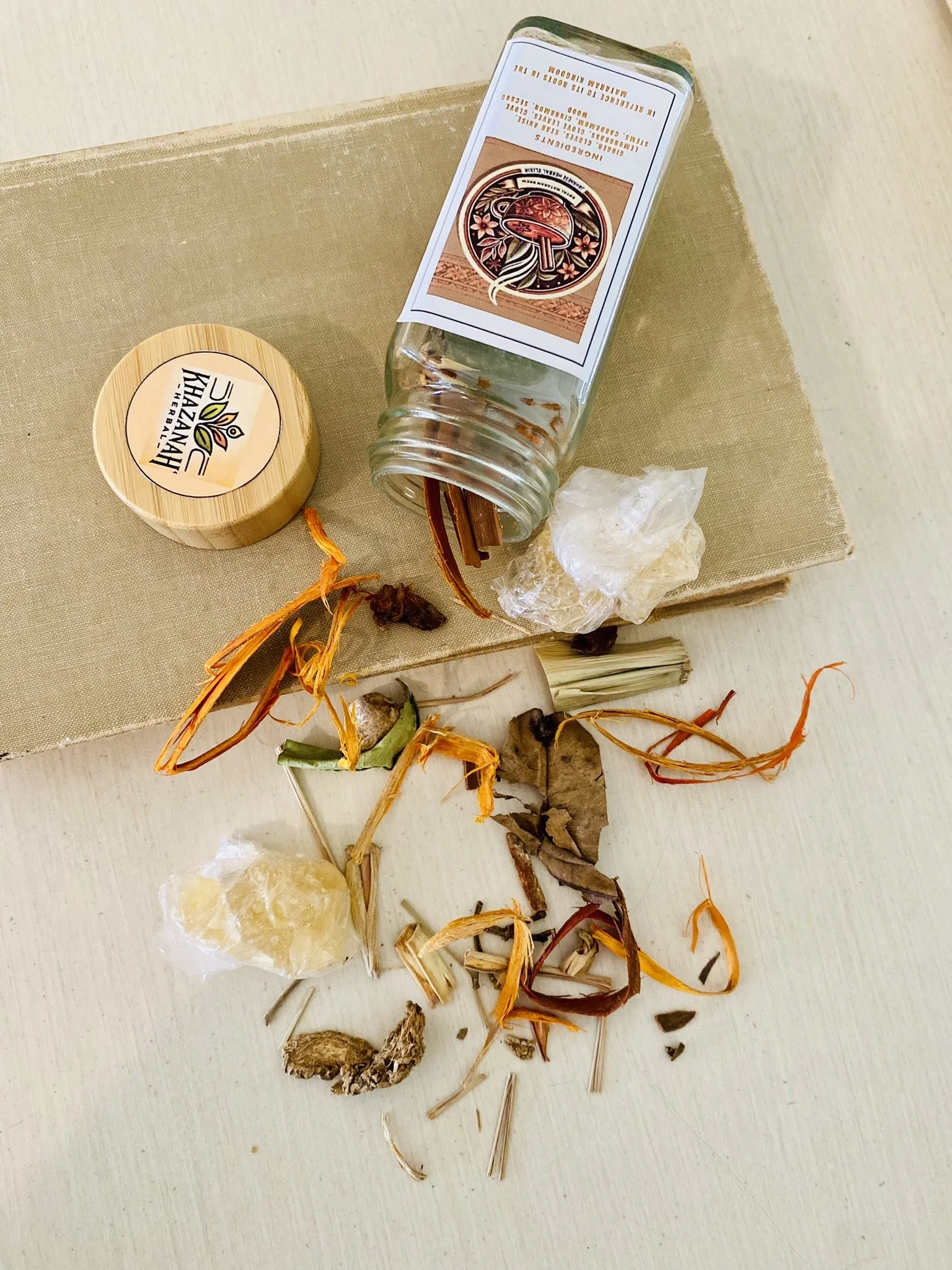 A collection of dried herbs and plant materials scattered on a white surface, with an open glass jar, a small wooden container labeled "Hazani" and a piece of beige cloth underneath.