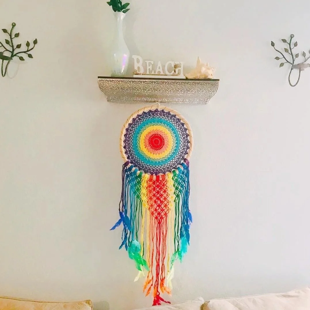 Colorful rainbow dreamcatcher hanging on a white wall with a shelf above it holding a white vase with green leaves, a decorative word 'BEACH', a shell, and a starfish.
