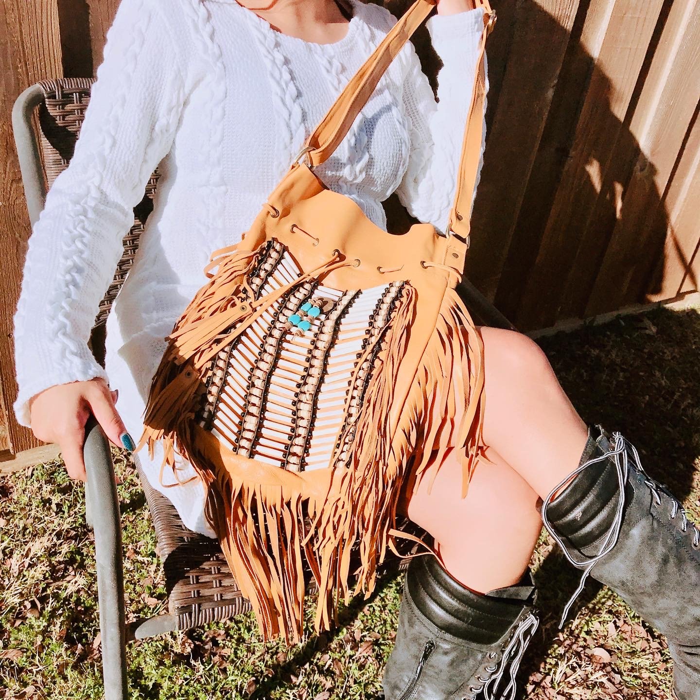 A person wearing black lace-up boots with heels, a white sweater, sitting on a wicker chair outdoors, holding a fringed leather bag with beads and turquoise jewelry.