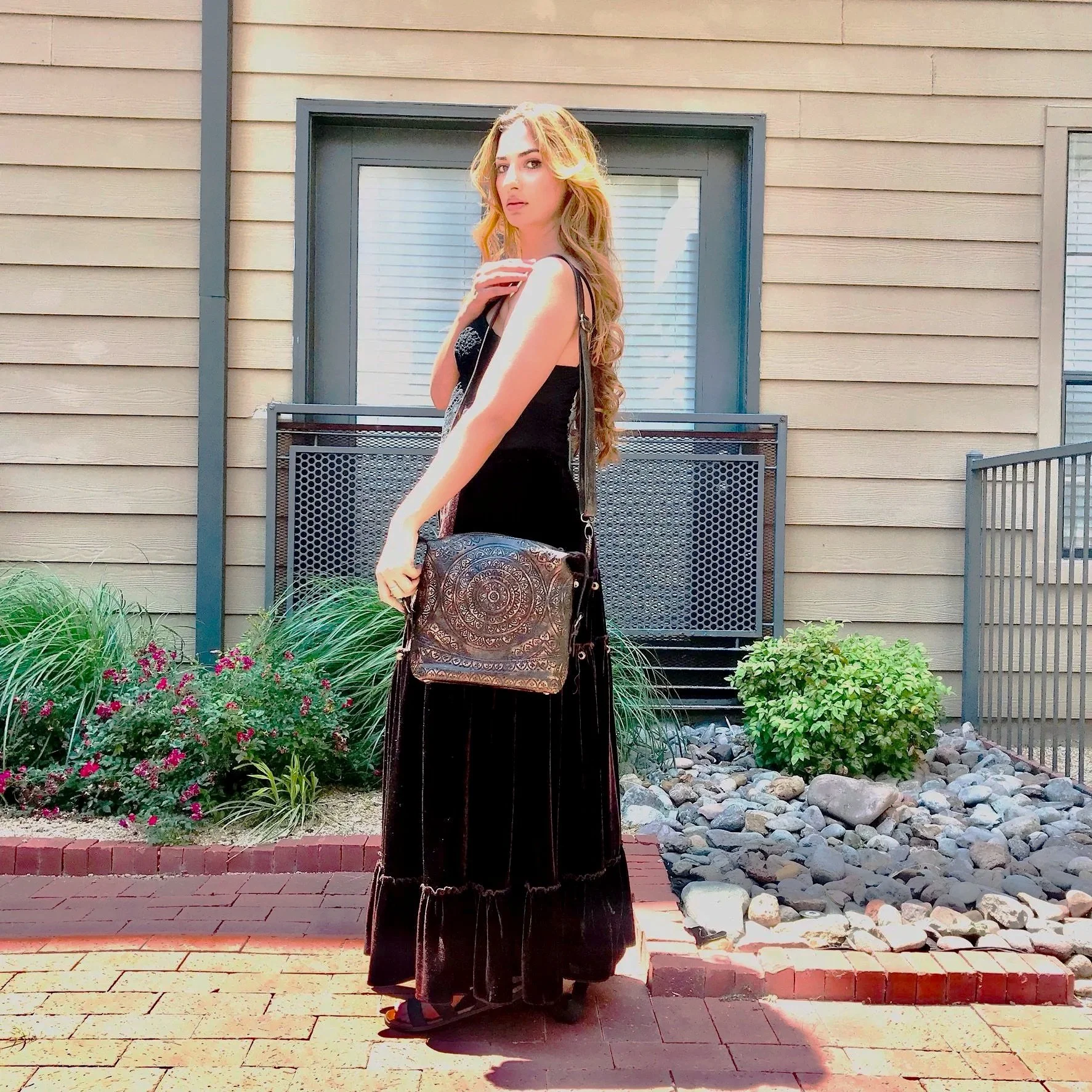 A woman with long, wavy hair dressed in a black sleeveless dress and black sandals standing on a brick sidewalk in front of a house with beige siding, a window, and a small garden with plants and rocks.