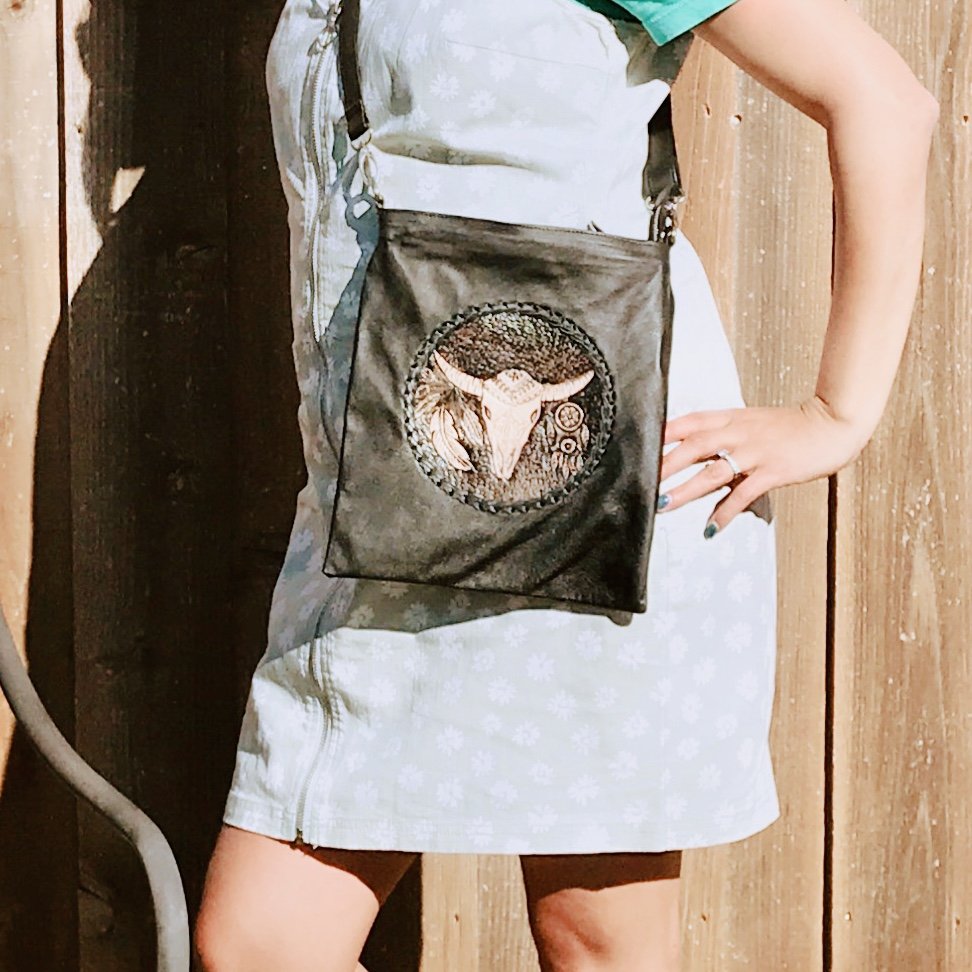 A person wearing a light-colored dress with a floral pattern, standing next to a wooden wall, carrying a black shoulder bag with a round cow head design on it.