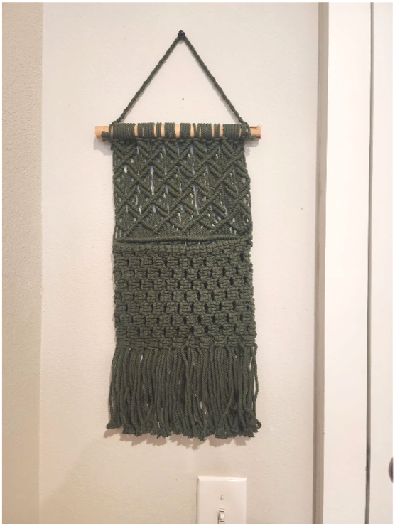 Green wall hanging with various knitted patterns and fringe at the bottom, suspended from a wooden rod with a black cord.