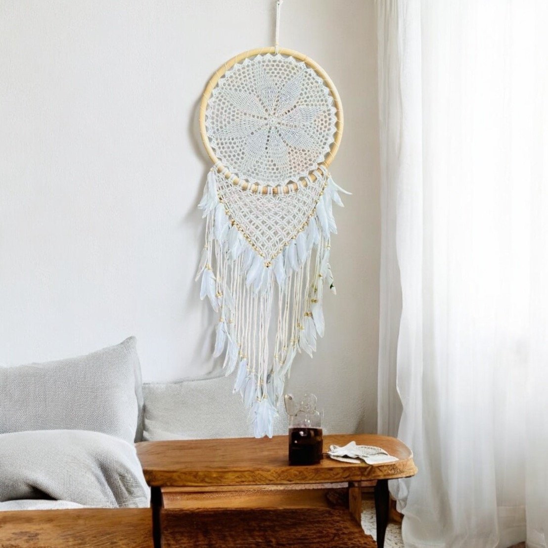 A bohemian-style bedroom decor featuring a large dreamcatcher with white feathers hanging on the wall above a wooden table, with a glass of dark beverage and a small stack of papers or napkins on the table.