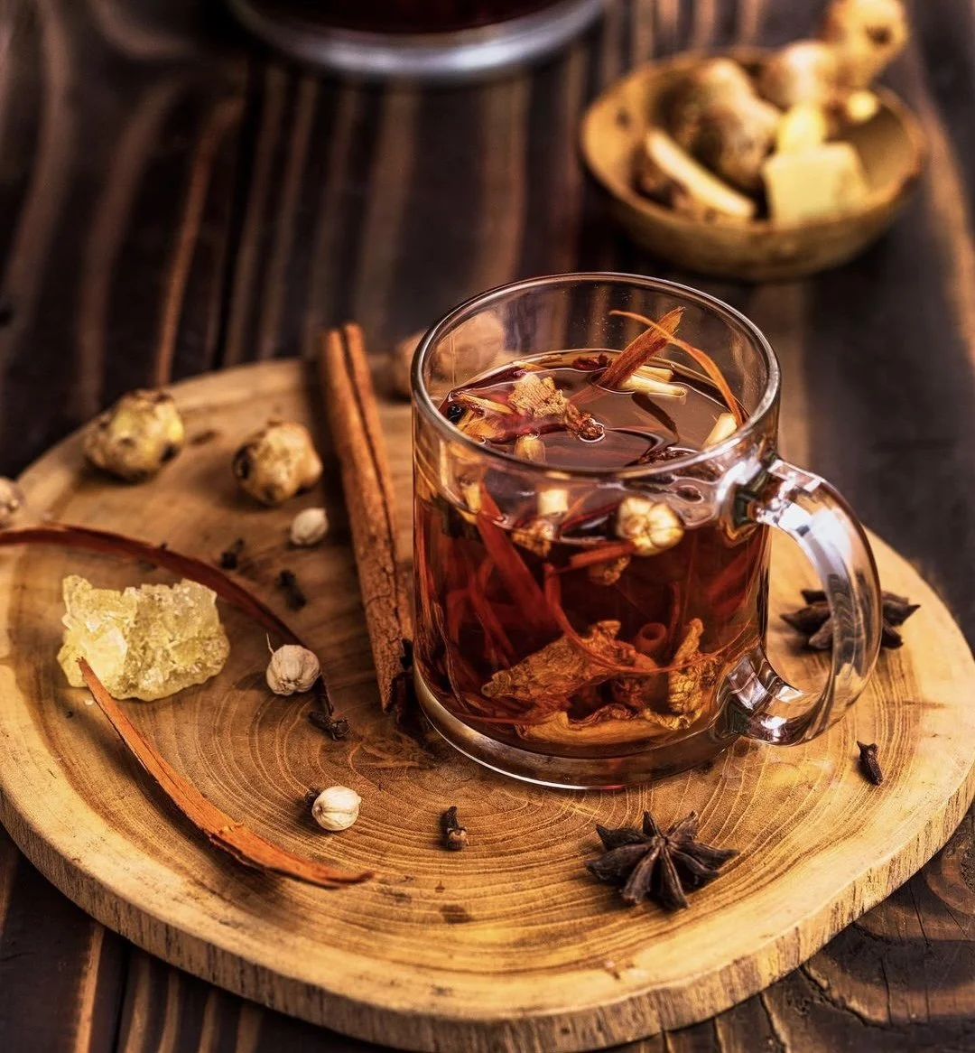A glass mug filled with hot herbal tea containing dried herbs, spices, and flowers, placed on a wooden tray with cinnamon sticks, star anise, whole garlic cloves, and ginger root, with a bowl of dried herbs and spices in the background.