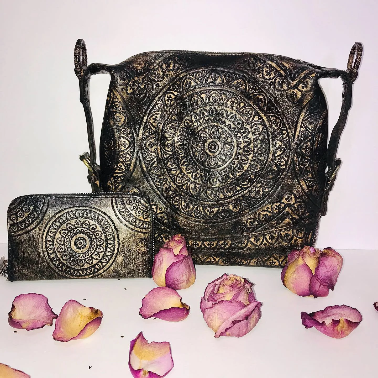 Matching black and gold jewelry set with intricate circular patterns, includes a large handbag and a smaller wallet, surrounded by scattered pink and yellow rose petals.