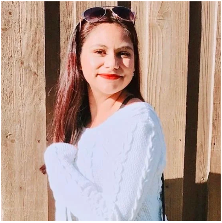A young woman with long brown hair wearing sunglasses on her head and red lipstick, smiling and looking at the camera, standing outdoors against a wooden fence.