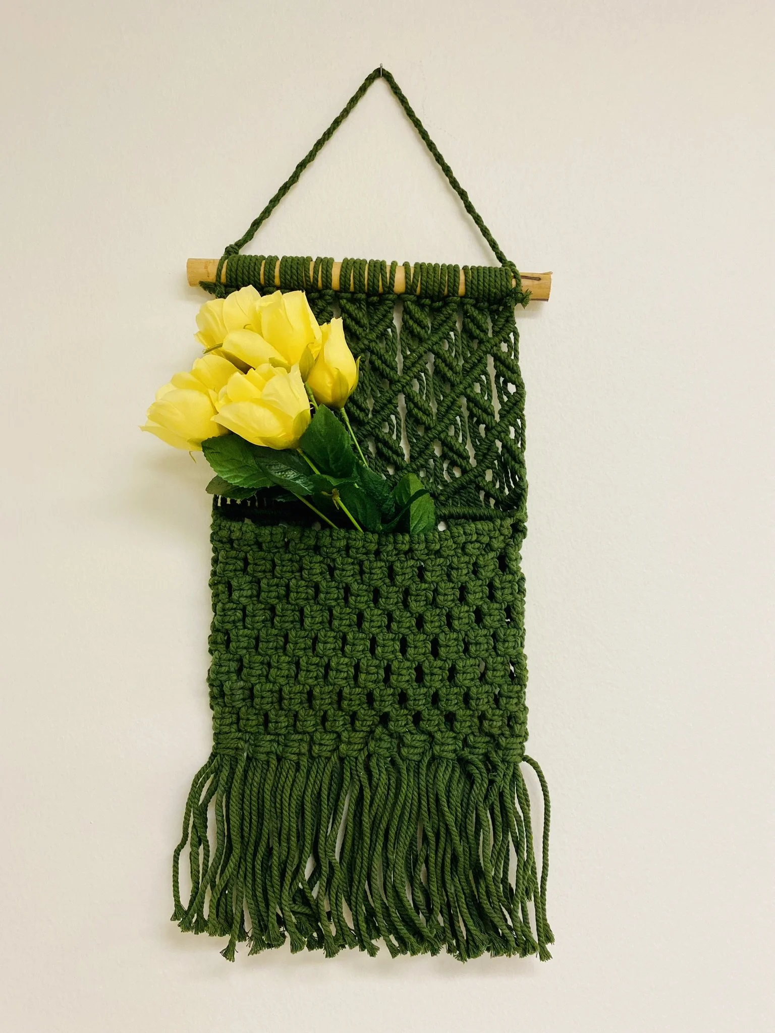 Green macrame wall hanging with fringes, small yellow roses with green leaves, and a wooden dowel at the top, hanging against a plain white wall.