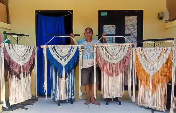 A smiling person standing behind a display of colorful, handmade macrame wall hangings hung on clothing racks indoors.