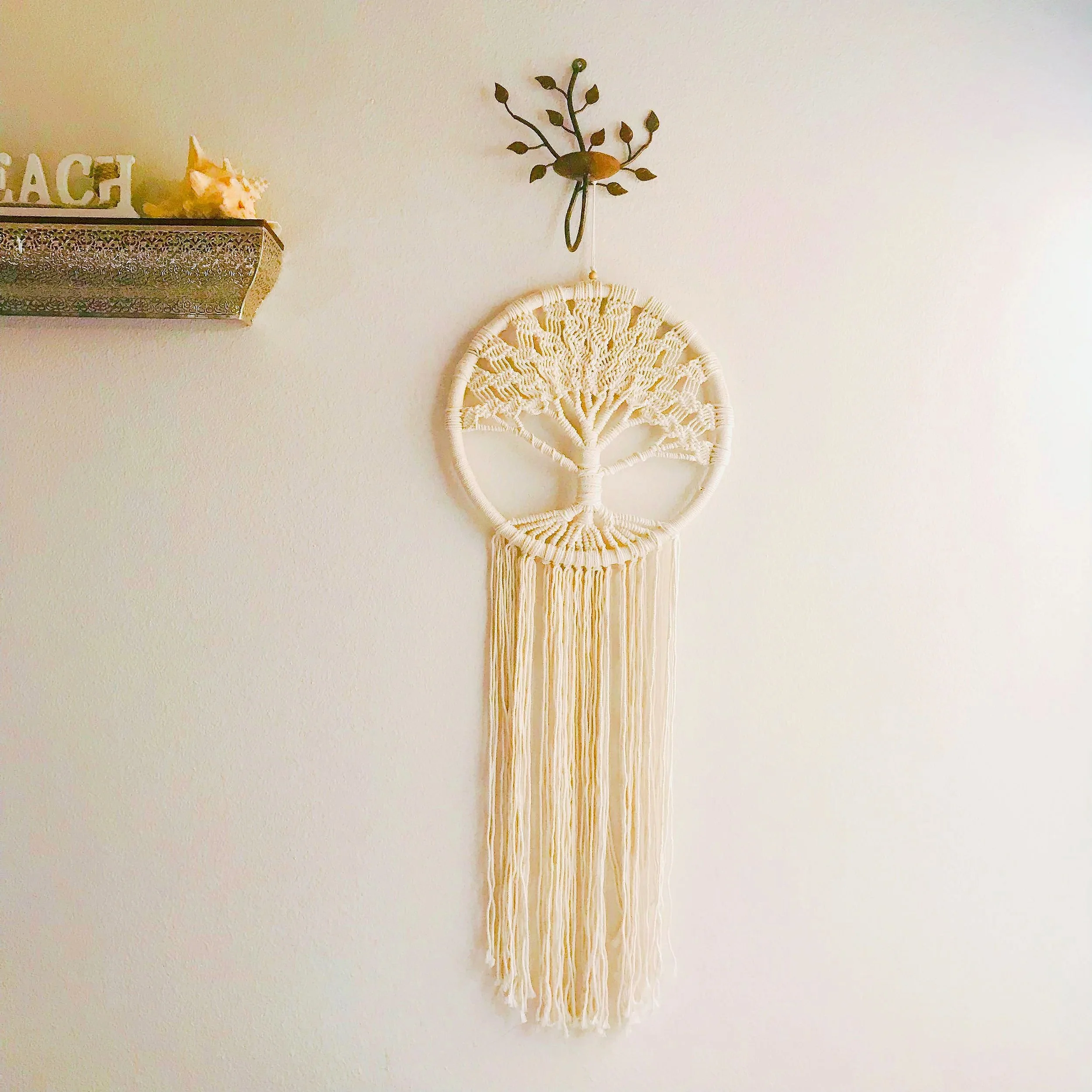 A cream-colored macramé wall hanging with a circular tree design and long hanging strands on a plain wall.