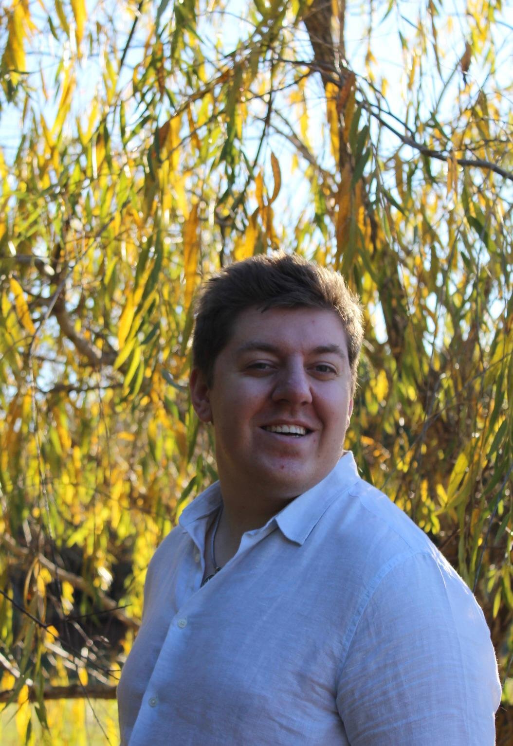 A young man with dark hair smiling in front of a background of yellow and green leafy trees during daylight.