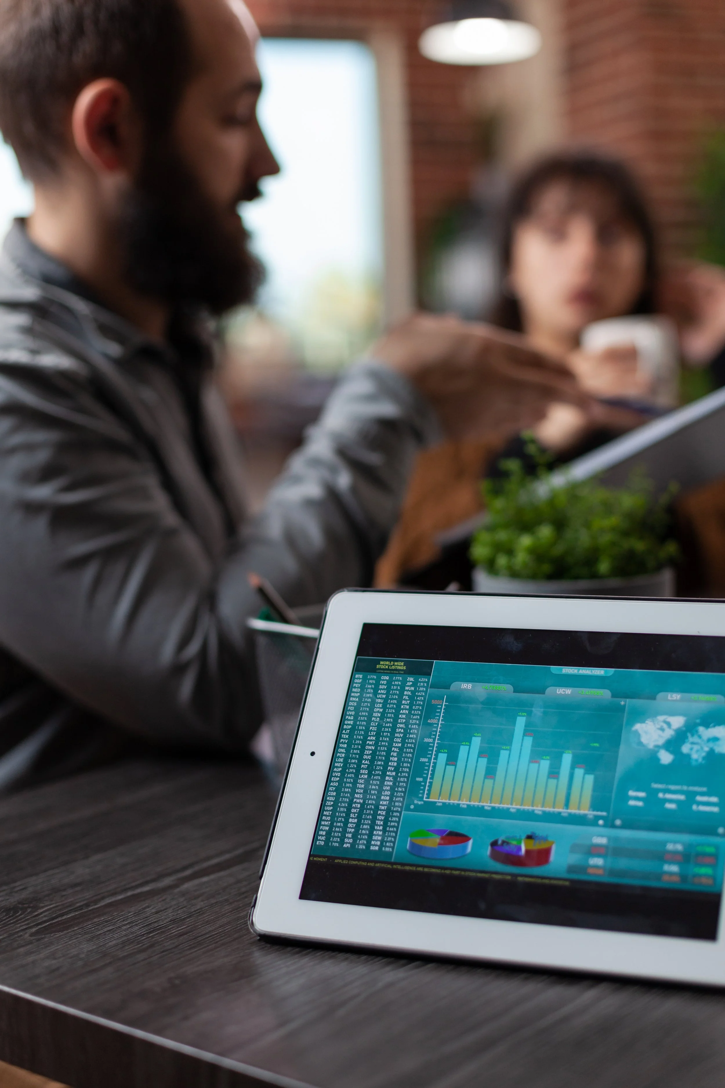Close-up of a tablet displaying colorful charts and graphs, on a table in a modern office with two blurred people working or having a meeting in the background.