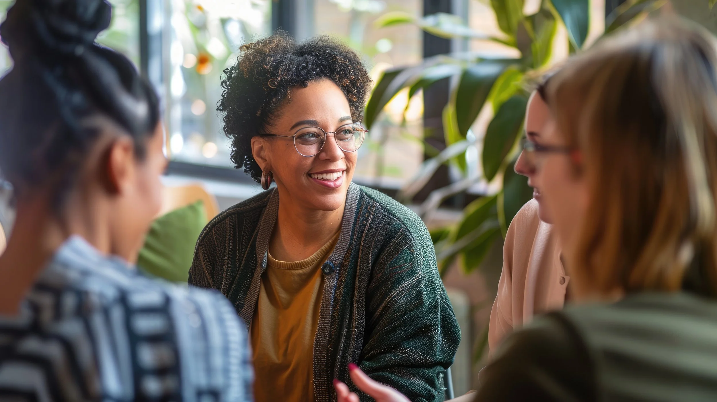 A diverse group of women engaged in a conversation indoors, with one woman smiling and wearing glasses, surrounded by large green plants and natural light.